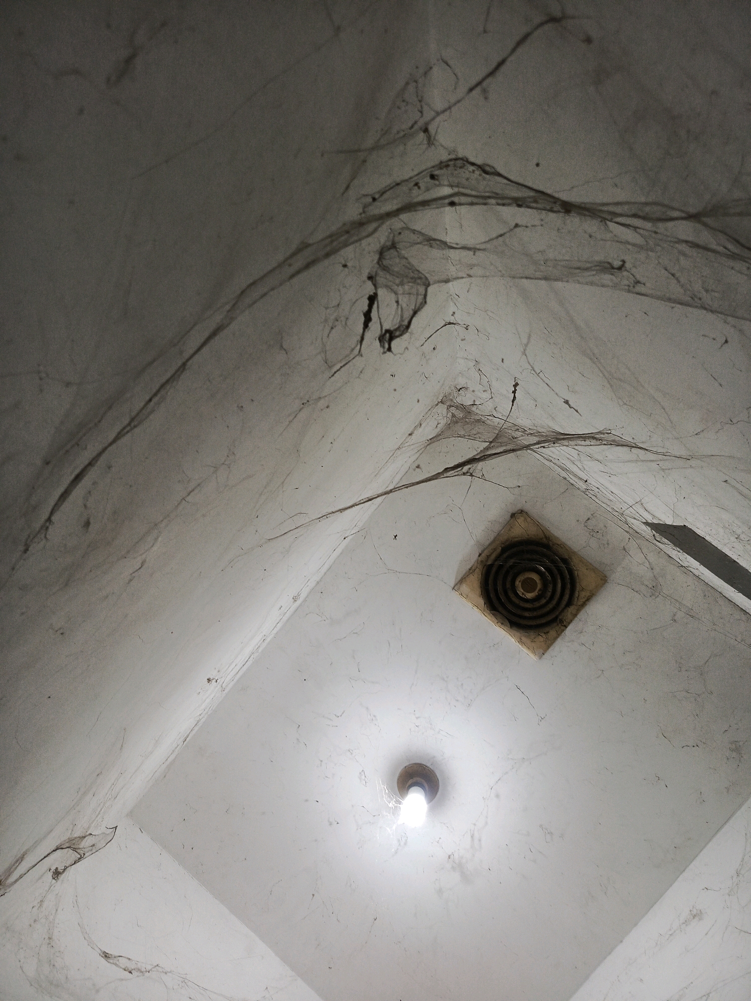 The image captures an interior ceiling area, likely within a neglected or disused building in Pamekasan, Indonesia. Cobwebs heavily adorn the stark white ceiling and corners, suggesting a prolonged period of disuse. A bare, lit lightbulb hangs centrally, casting a soft glow. To the right of the lightbulb, a square vent with concentric circular openings is visible, also covered in a layer of dust and cobwebs. The overall impression is one of abandonment and decay.