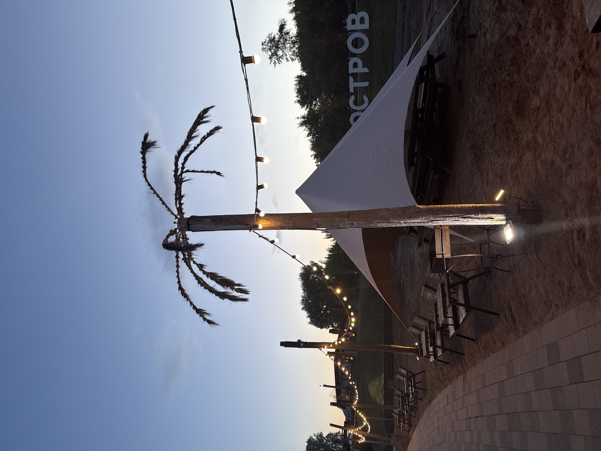 The image depicts an outdoor recreational area at dusk in Votkinsk, Russia. The scene features sandy ground, an illuminated paved pathway, and various artificial lighting installations.
In the foreground, a curvilinear path constructed from interlocking gray and reddish-brown pavers extends along the left edge. Adjacent to the path, the ground transitions to sand, upon which multiple sets of dark wooden tables and chairs are arranged. Near the front right, a portable metal brazier or charcoal grill on an ornate stand is positioned next to a table.
Several wooden poles, stained dark, are vertically erected within the sandy area. These poles are designed to resemble palm trees, featuring crown-like structures made of dried, bristly material. Strands of warm-toned, round light bulbs are strung between these poles, providing ambient illumination. Some poles also have ground-level or pole-mounted spotlights illuminating their bases and the surrounding sand.
Towards the right, a large, light-colored fabric canopy or stretch tent covers a section of the sandy area, providing shelter. Behind this tent, a dense line of dark green trees forms a natural backdrop. On a grassy slope behind the trees and partially visible through the tent, large white three-dimensional letters spell "ОСТРОВ" (OSTROV), which translates to "Island."
The sky transitions from a deep blue at the zenith to a lighter, yellowish-orange hue near the horizon, indicative of late evening or early morning. A few faint clouds are visible. No people are present in the image, and no specific actions or interactions are depicted. The overall environment suggests a prepared outdoor dining or leisure space.