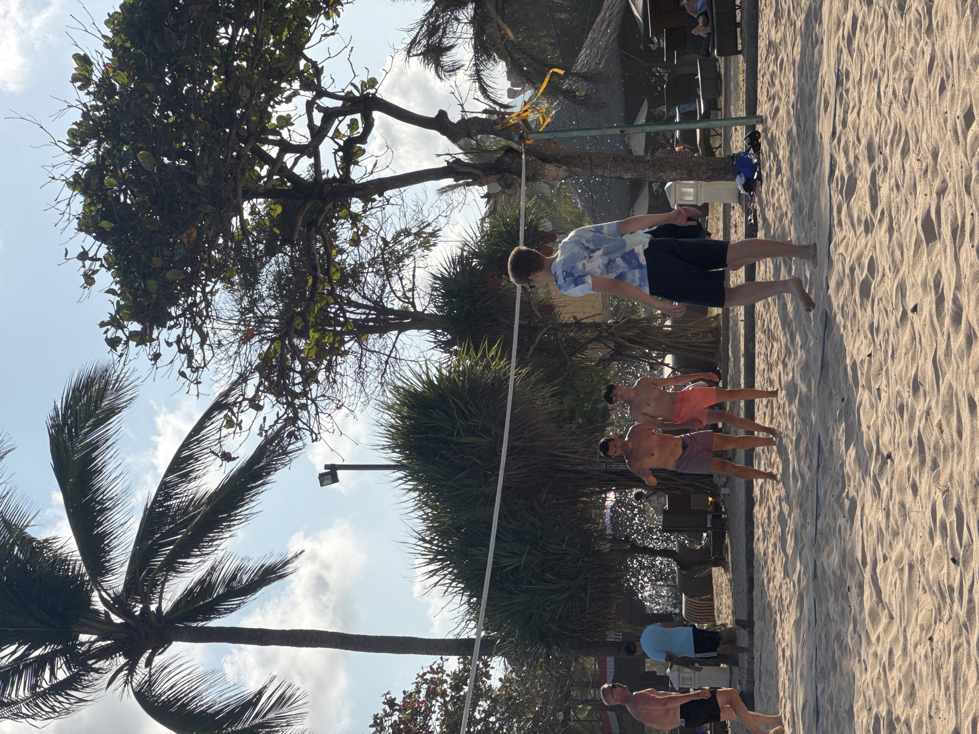 Four young men are playing volleyball on a sandy beach in Kuta Selatan, Indonesia. Two men are standing near the net, facing away from the camera, while two others are positioned further down the beach. The men are all wearing shorts, and one is shirtless. Palm trees and lush vegetation surround the beach. The sky is partly cloudy, and the sun is shining. A volleyball net is set up across the court. In the background, there are thatched-roof structures, lounge chairs, and other beachgoers. The scene is vibrant and energetic, capturing the essence of a tropical beach vacation.