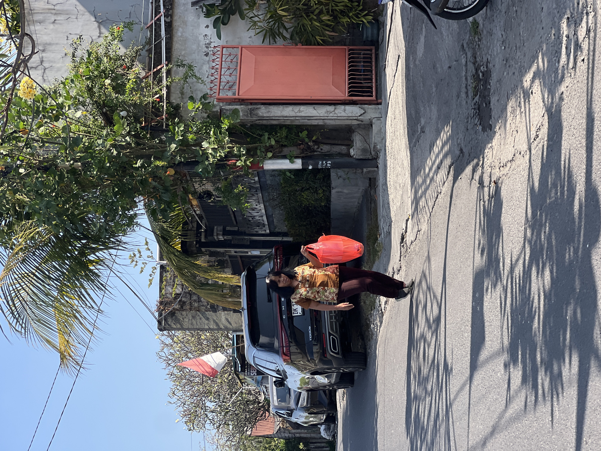 A woman walks along a paved street in Denpasar, Indonesia, on a sunny day. She is wearing a brown and orange floral print blouse and dark maroon pants. In her right hand, she carries a bright red plastic bag. To her left, a black SUV is parked on the side of the road. Behind the SUV, a red and white Indonesian flag is visible. The street is lined with buildings and vegetation, including palm trees and other lush green plants. The sun casts long shadows on the asphalt, creating a dappled pattern. A red metal gate is visible to the right of the woman.