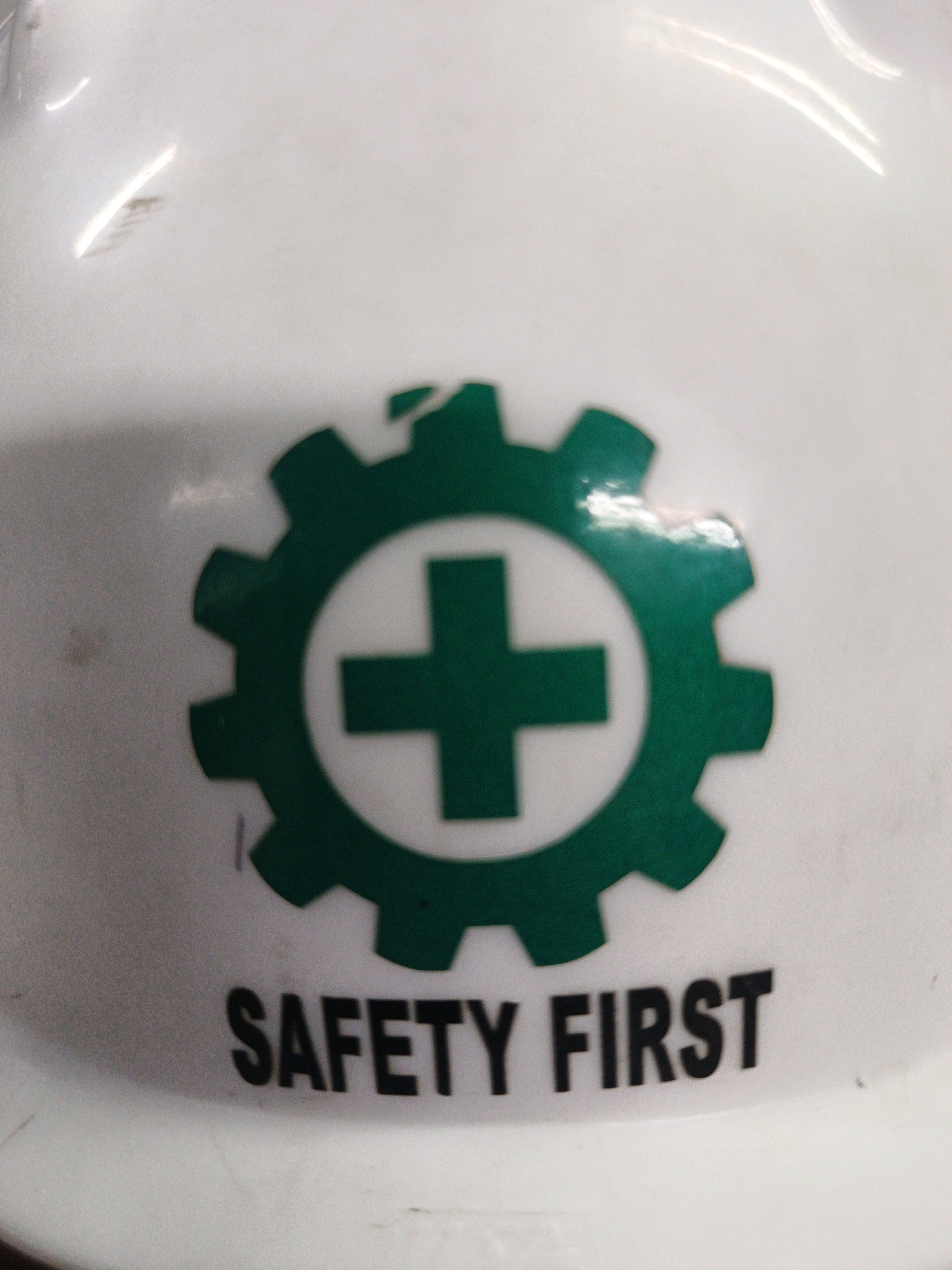A white hard hat is shown with a green gear-shaped logo containing a white cross in the center. The words "SAFETY FIRST" are printed in black text below the logo. The image appears to be taken in Ngoro, Indonesia, likely in an industrial setting given the presence of a safety helmet. The background is blurred and light-colored, suggesting an indoor environment. No people are visible in the image.