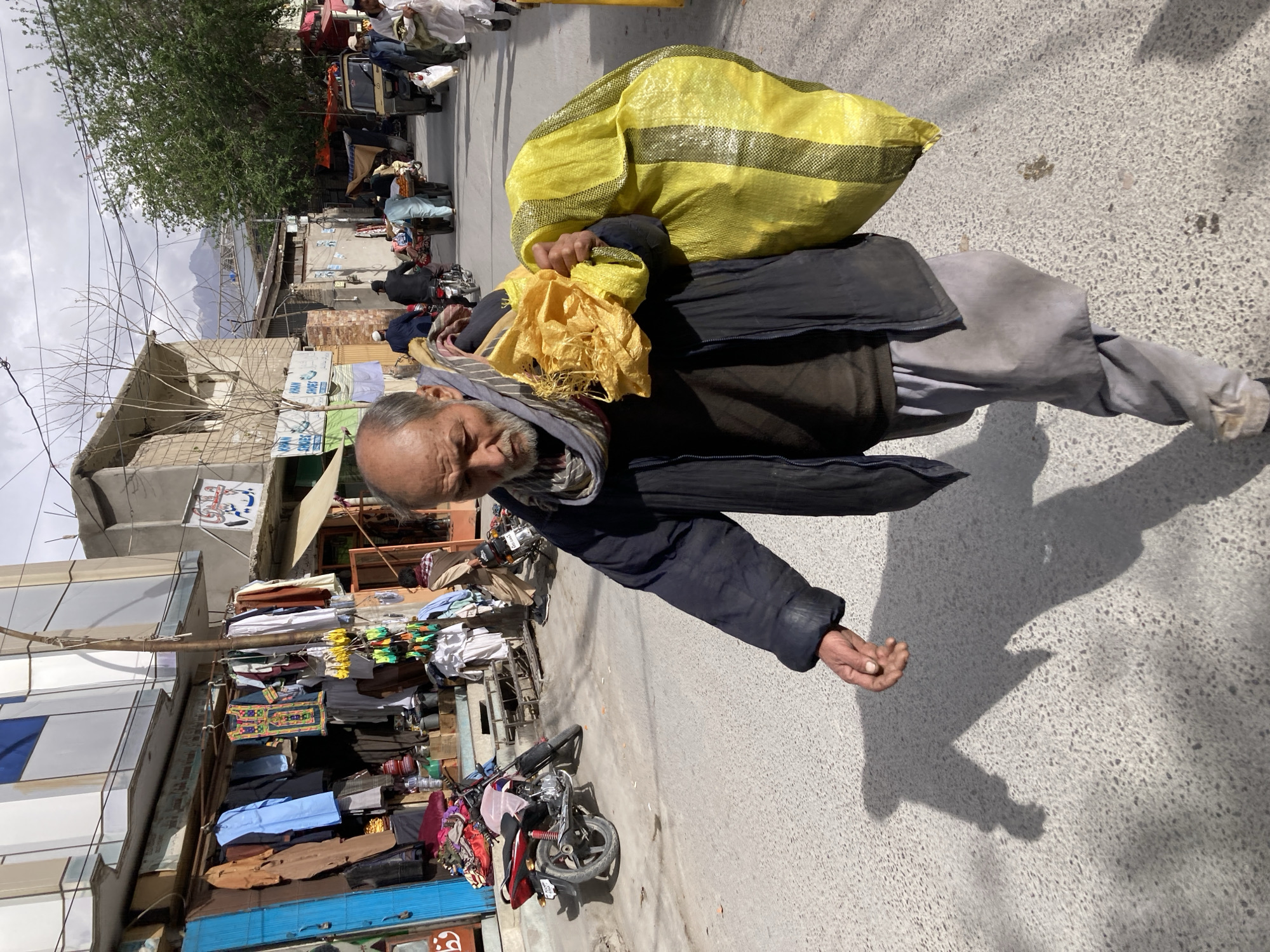 In Quetta City Tehsil, Pakistan, an elderly man walks along a paved street carrying a large yellow bag over his shoulder. The street is lined with shops and stalls selling clothing and other goods. A motorcycle is parked nearby. Other pedestrians and vehicles can be seen in the distance. The sky is overcast.