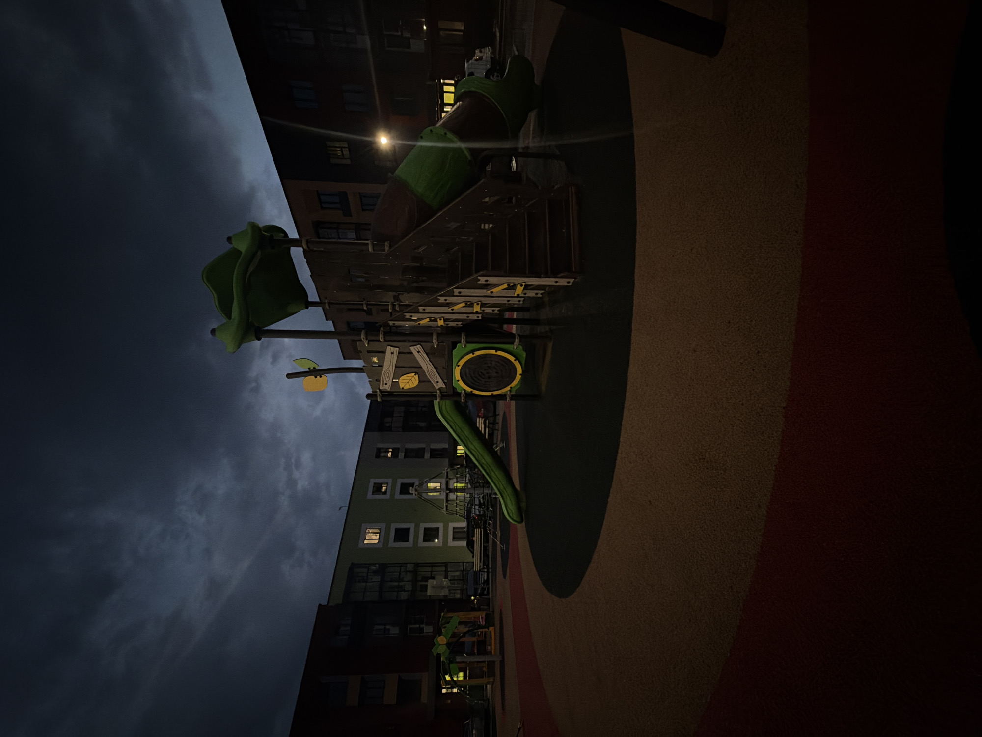 The image shows a children's playground located in the city of Фролы, Russia, at dusk or night. The playground features a green tube slide, a set of wooden stairs leading up to it, and a climbing net structure. The ground of the playground is covered with dark, textured material, with a circular section in the foreground that is a mix of red and yellow. In the background, apartment buildings with lit windows are visible. The sky is dark with heavy clouds. There are no people or animals present in the image.