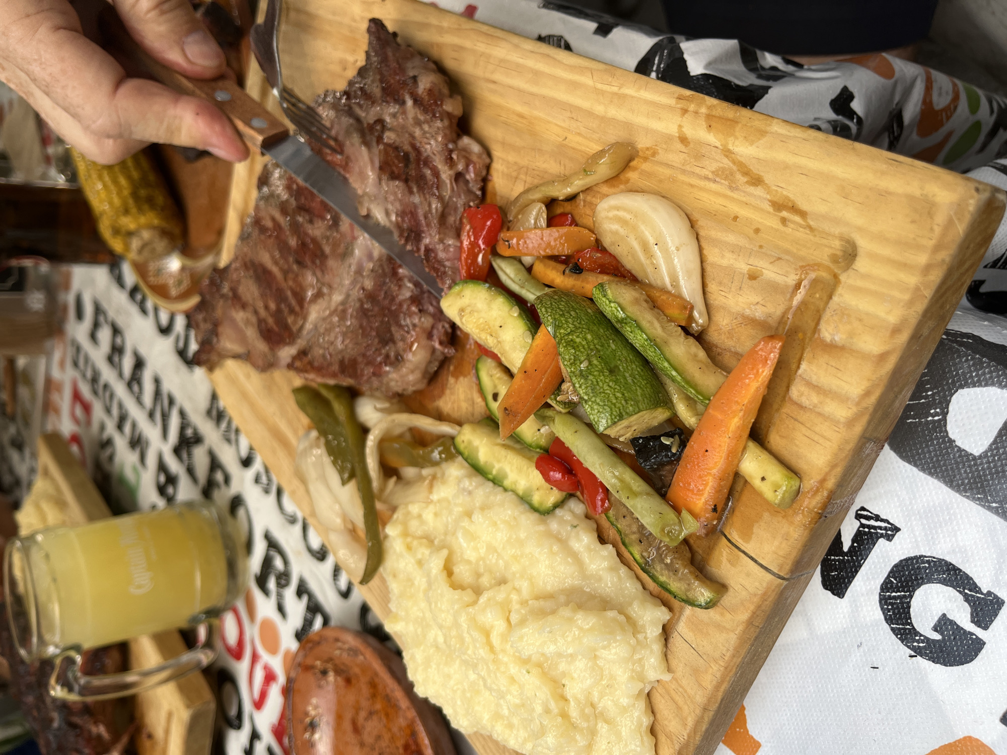 A wooden cutting board holds a grilled steak, sauteed vegetables (zucchini, carrots, peppers, and onions), and mashed potatoes. A person is using a fork and knife to cut the steak. A glass of yellow liquid sits beside the cutting board. The setting appears to be a table in a restaurant or home. The environment contains tablecloths with bold prints. It is difficult to determine the location.