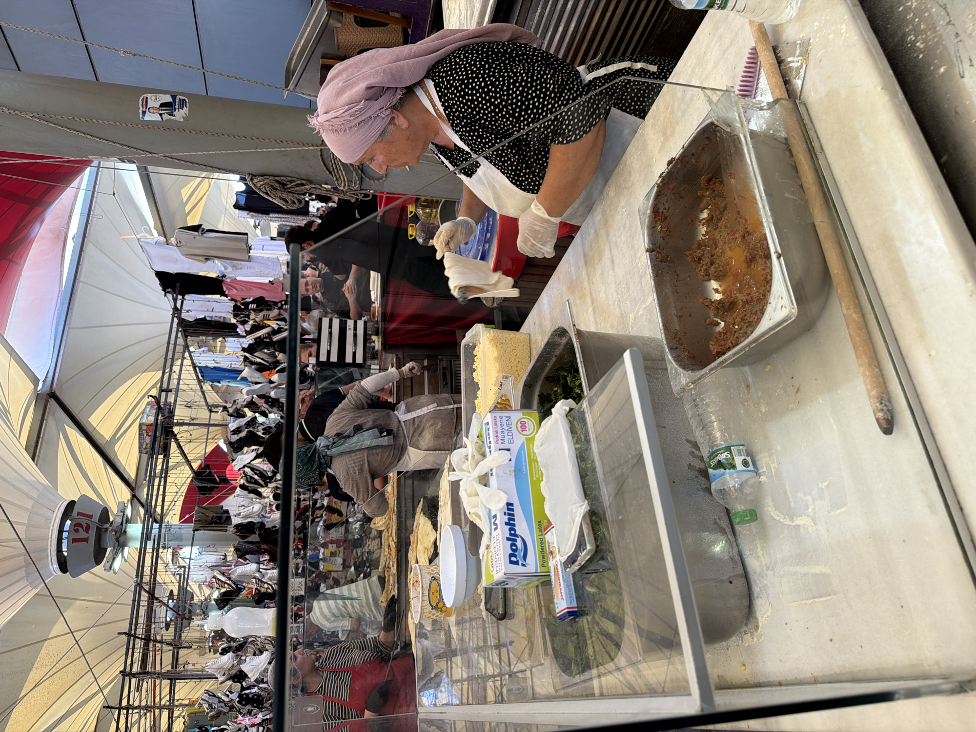 The image depicts an indoor market scene, likely in Kadıköy, Turkey, focusing on a food preparation area. In the foreground, a woman wearing a light pink headscarf and a black and white polka dot dress, with white gloves and apron, is seen holding a piece of dough. She is standing behind a glass counter, which separates her from the viewer. To her right, a metal tray contains a mixture of ground meat and spices. In front of the counter, several food preparation items are visible, including a box of "Dolphin" powdered latex gloves, a white bowl, and a plastic water bottle.
The background reveals more of the market, with stalls displaying various items hanging from the ceiling, including clothing and footwear. Other individuals are visible in the background, engaged in activities related to food preparation or sales. The overall environment appears to be a bustling, open-air market under a large canopy. A sign with "121" is visible hanging above.