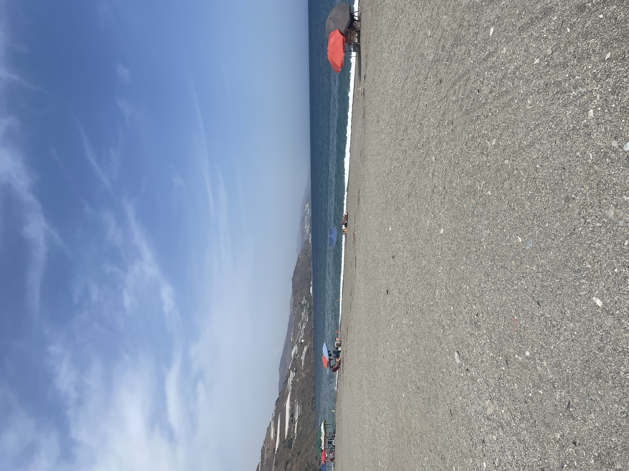 The image depicts a pebble beach in Lújar, Spain, under a bright blue sky with scattered white clouds. The sea is calm with gentle waves breaking on the shore. The beach is sparsely populated with several beach umbrellas providing shade for a few individuals.
In the foreground, the dark, pebbly texture of the beach dominates the lower half of the frame. A few scattered, lighter-colored stones and debris are visible.
Moving towards the mid-ground, the transition to the sea is marked by a white line of breaking waves. Several beach umbrellas are set up. On the left, a few umbrellas are clustered together, with people seated underneath. Further to the right, a red and a grey umbrella are prominent, shading a table and chairs. A few individuals are seated or standing near these umbrellas. In the sea, a few people can be seen swimming or wading.
The background features the coastline of Lújar, characterized by a steep, mountainous terrain. The hills are covered in dark vegetation and numerous white greenhouses, indicative of agricultural activity. The horizon line clearly separates the sea from the sky. The overall scene suggests a tranquil day at the beach.