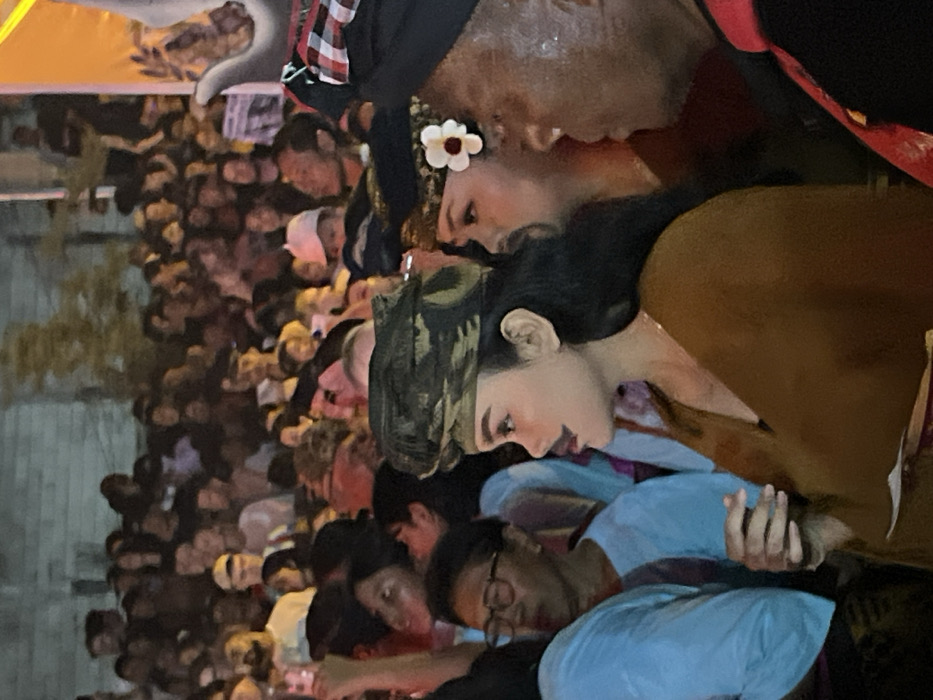 The image depicts a crowded outdoor nighttime scene in Munggu, Indonesia. In the foreground, a woman with dark hair wears a camouflage-patterned headwrap, a brown garment, and has a painted mustache and red lipstick; she holds a small white object in her right hand and looks toward the right. Behind her and slightly to the right, another woman is partially visible, wearing a dark headwrap with a white flower embellishment. To the far right, a man with dark skin, wearing a traditional red and white checkered Balinese udeng and a black and red garment, is partially visible, facing left. To the left, a woman wearing glasses and a light blue top observes the scene. The background is filled with a dense, out-of-focus crowd of people under artificial lighting, suggesting a public event or performance.