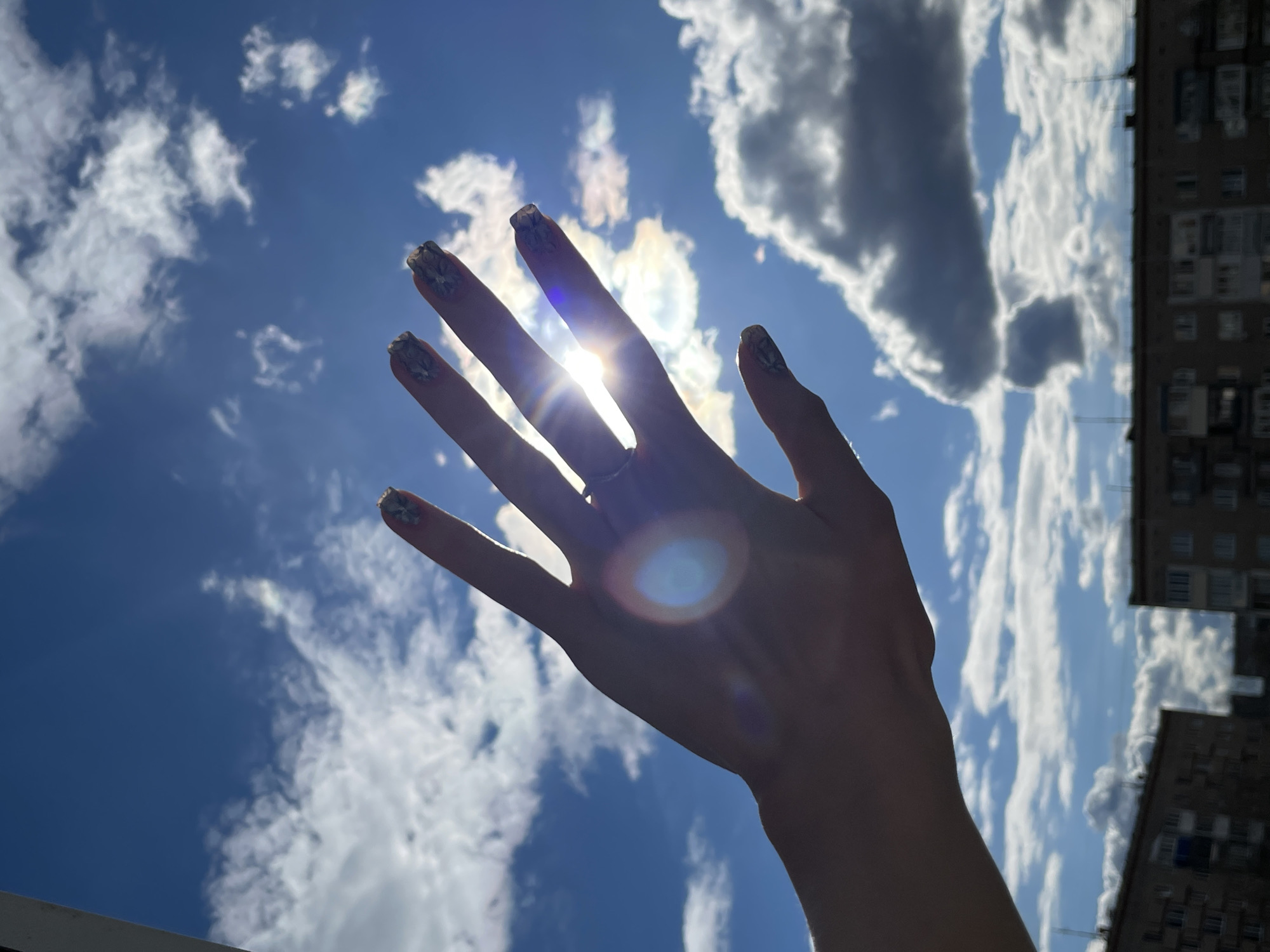 The image presents a bright outdoor scene on a sunny day in Izhevsk, Russia. A human hand, likely female due to its slender appearance and decorative fingernails, is raised against a vibrant blue sky filled with scattered white cumulus clouds. The sun is positioned directly behind the hand, casting it in silhouette and creating a prominent lens flare that emanates from between the fingers and forms a distinct circular glow on the palm. The nails are painted with an intricate, possibly floral or abstract, pattern in light tones. A ring is visible on one of the fingers.
On the far right side of the frame, a tall, multi-story residential building, typical of urban or suburban architecture, is partially visible and silhouetted against the bright sky. The building shows multiple windows and what appear to be antennas on its roofline. The overall lighting and clear sky indicate daytime, likely mid-day or early afternoon, with strong sunlight. There is no visible text in the image.