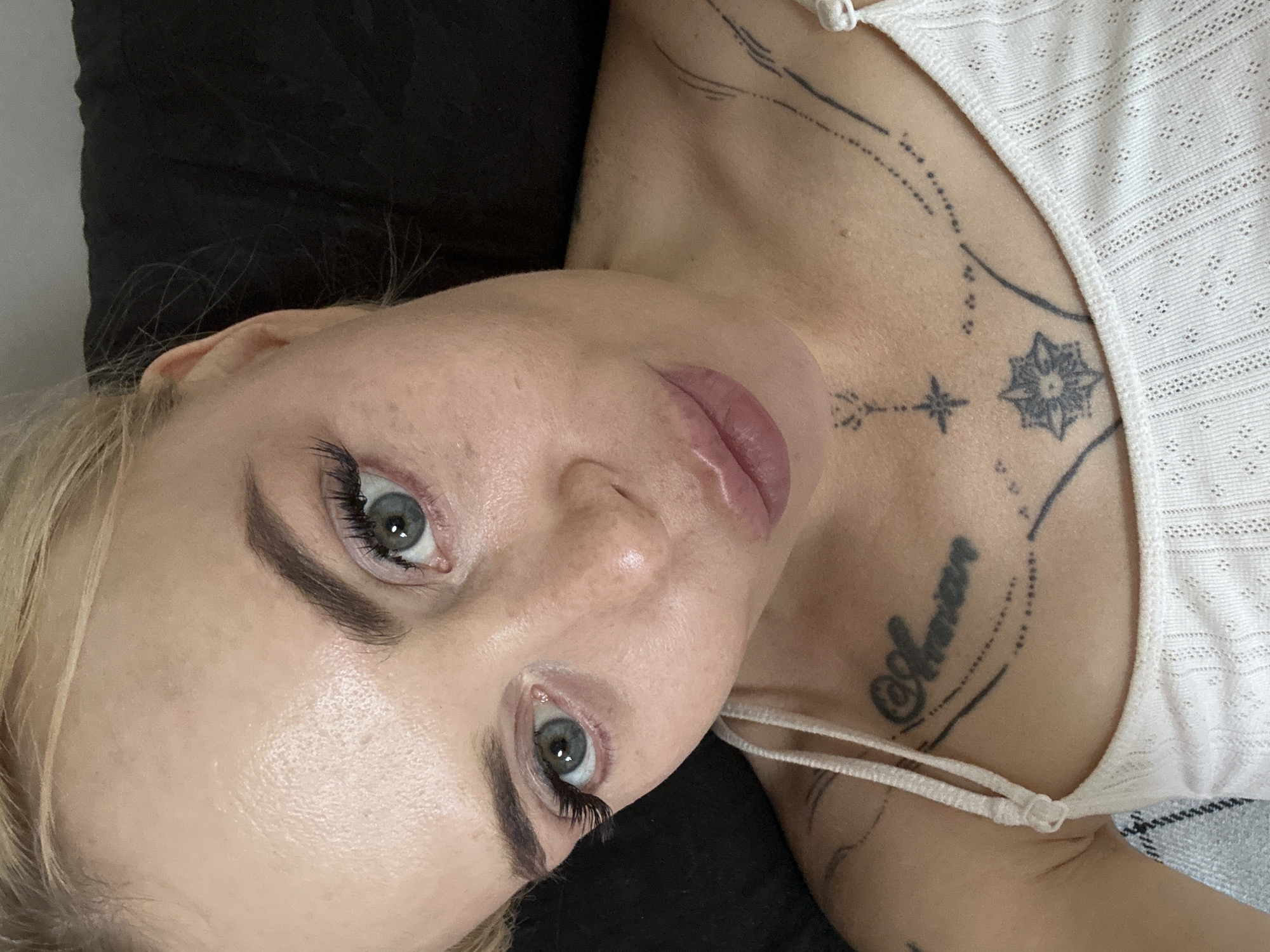 A woman with blonde hair and green eyes is shown from the chest up, looking directly at the camera. She is wearing a white tank top with thin straps. She has a tattoo on her chest that appears to say "Marian" and other decorative tattoos on her upper chest and shoulder. The background is dark and out of focus.