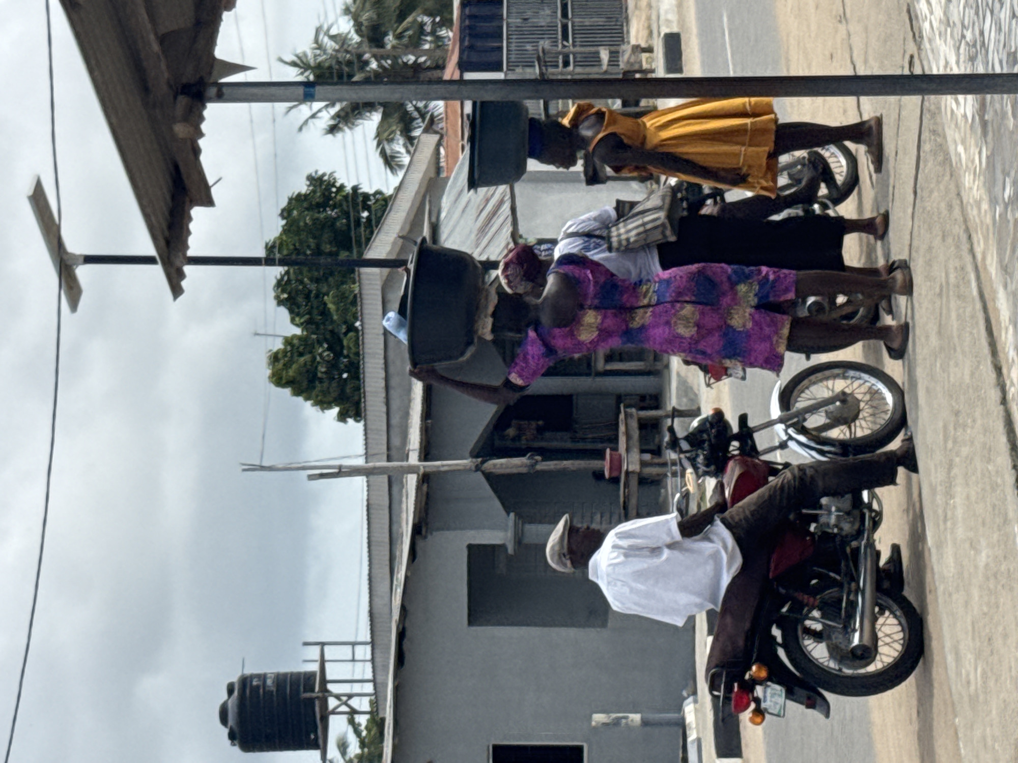 Captured in the city of Badagry, Nigeria, the image shows a street scene with people and motorbikes. A man wearing a white shirt and a cap is sitting on a red motorbike parked on the right side of the road. Close by, two women are walking alongside the road, each carrying large containers on their heads. One is wearing a purple patterned dress, the other a white top. A third woman in a yellow dress is walking further away. Houses with corrugated iron roofs line the street, with a water tank visible behind one of the buildings. Overhead, wires and a metal post are stretched across the scene. The sky is overcast.