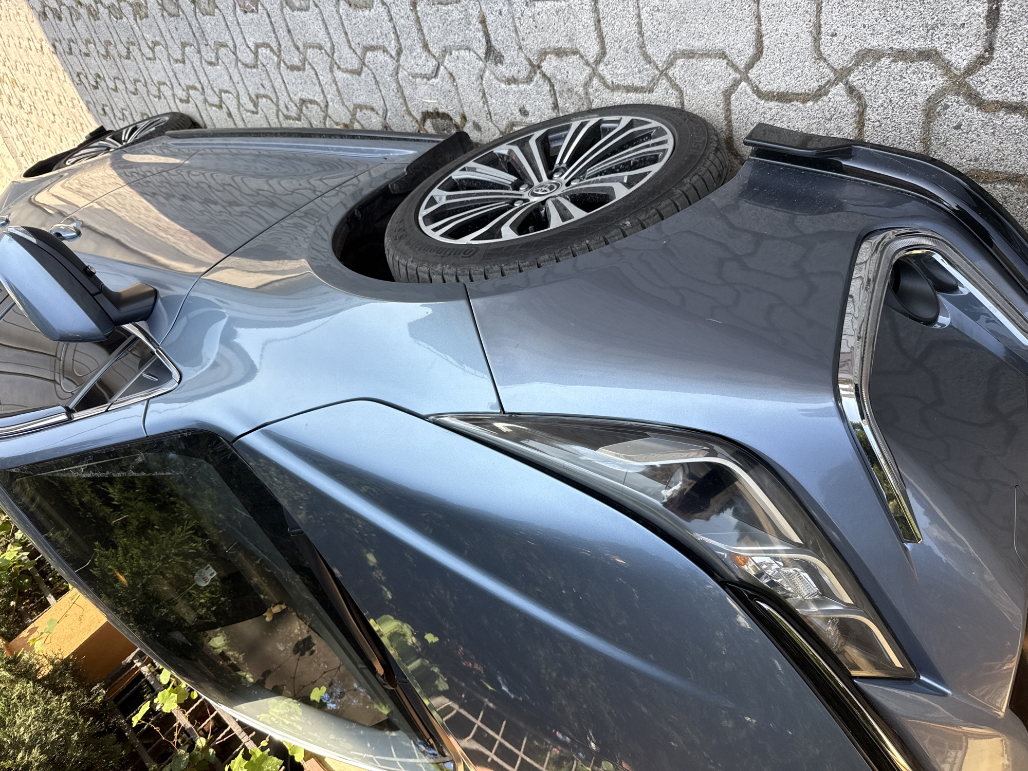 The image shows the front side of a blue car parked on a cobblestone surface in Sancaktepe, Turkey. The car's headlight, fender, and wheel are visible, along with a side mirror. The car is angled, with the front left of the vehicle closest to the camera. The ground is paved with light-colored interlocking cobblestones. Some greenery is visible in the background, suggesting an outdoor setting. The lighting indicates it is daytime, with shadows cast on the ground and the car's surface.