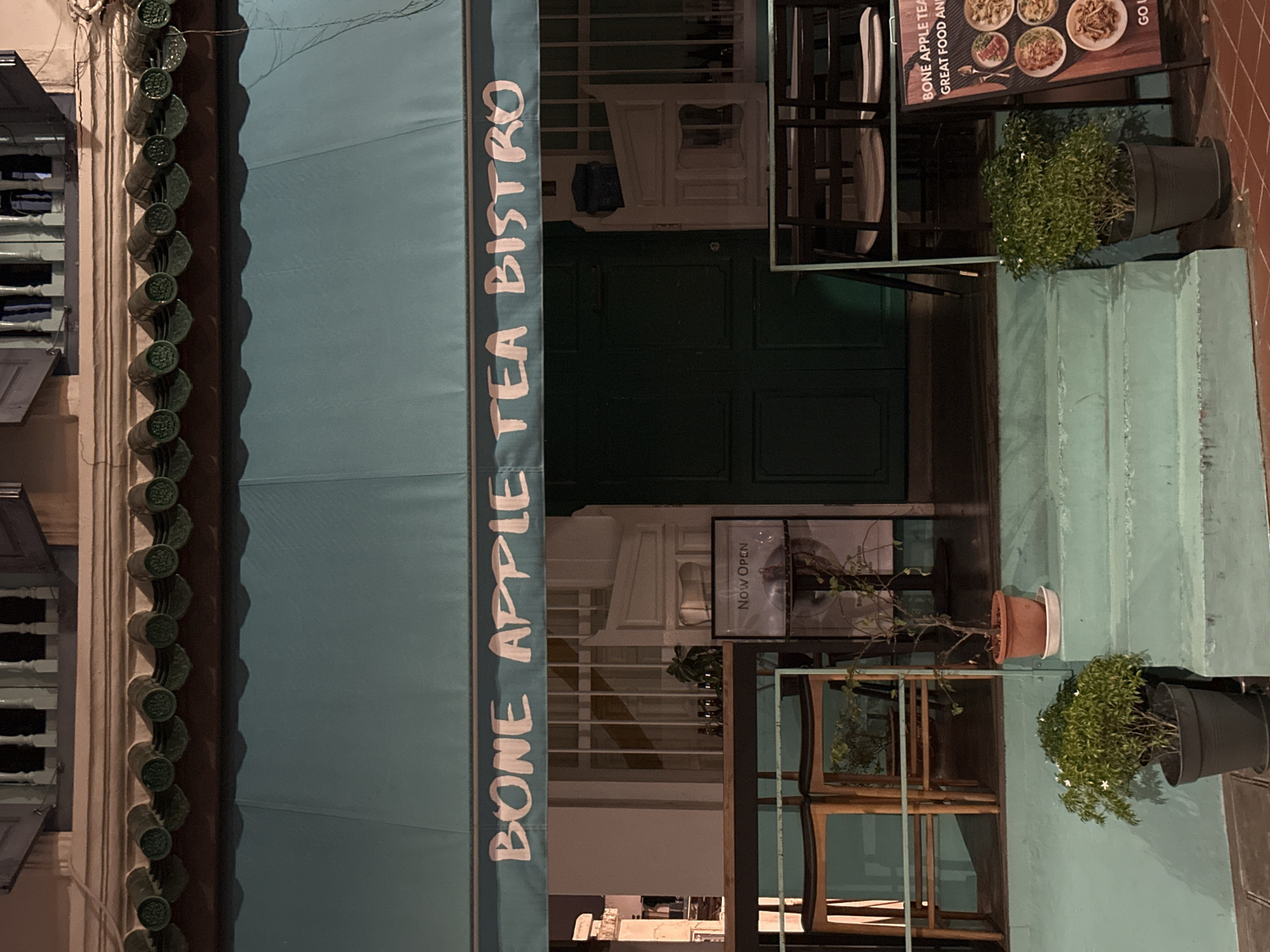 The image shows the exterior of a bistro named "BONE APPLE TEA BISTRO" in Singapore. The bistro has a teal-colored awning with the name written in white, cursive font. Below the awning, there is a dark green door and several windows with white grills. A set of teal steps leads up to the entrance, with potted plants on either side. To the right of the entrance, a sign displays images of food and the text "BONE APPLE TEA BISTRO GREAT FOOD AND DRINKS". The building appears to be an older shophouse, typical of areas like Chinatown or Kampong Glam in Singapore. The lighting suggests it is nighttime, and the scene is quiet and deserted.