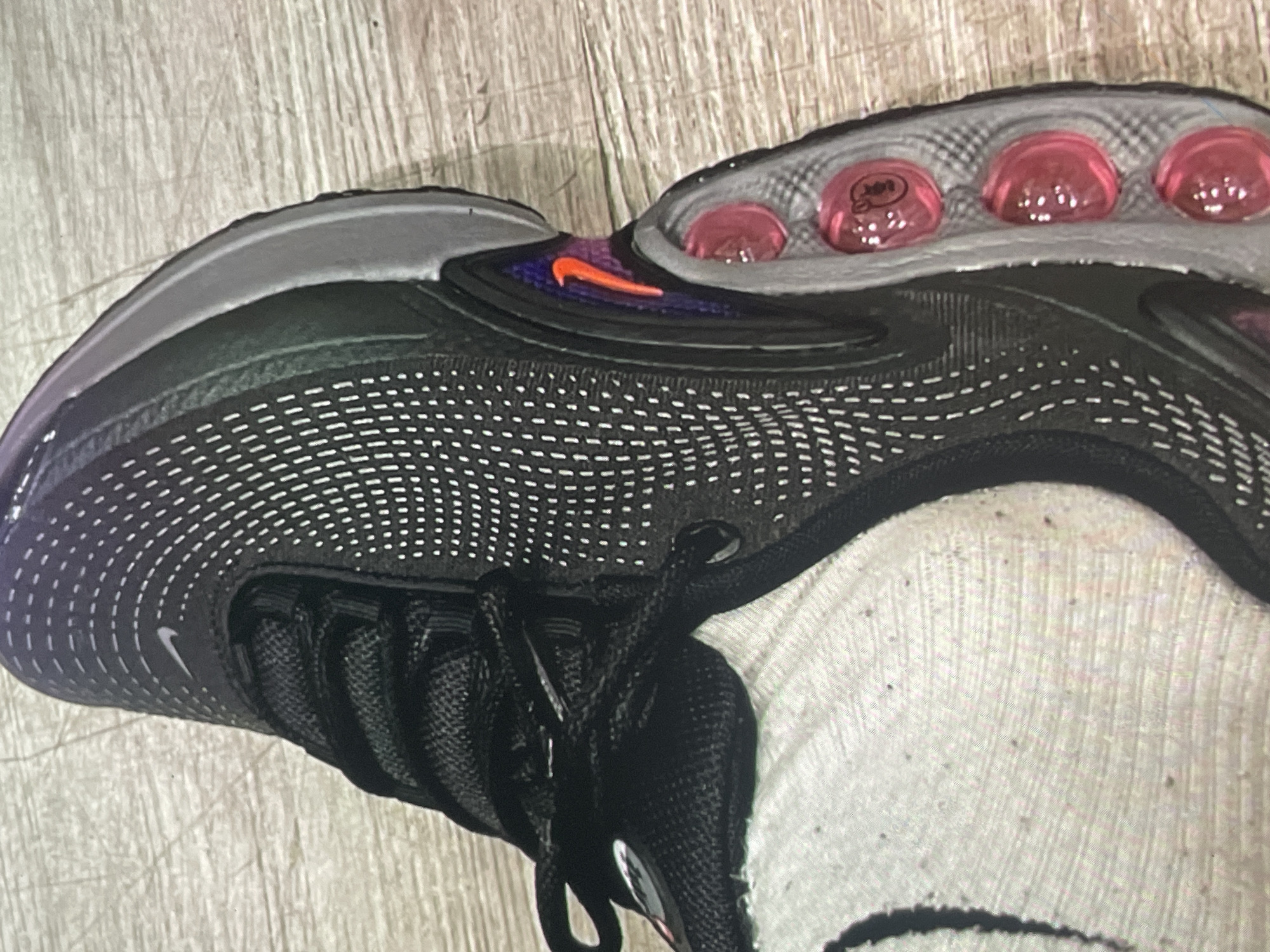 The image shows a close-up of the top and side of a Nike Air Max shoe. The shoe features a predominantly black upper with a white dotted pattern and black laces. The sole has pink air bubbles, and there is an orange Nike swoosh logo near the back. The shoe appears to be on a light-colored surface. The location of Harmanli, Bulgaria, is not evident within the image.