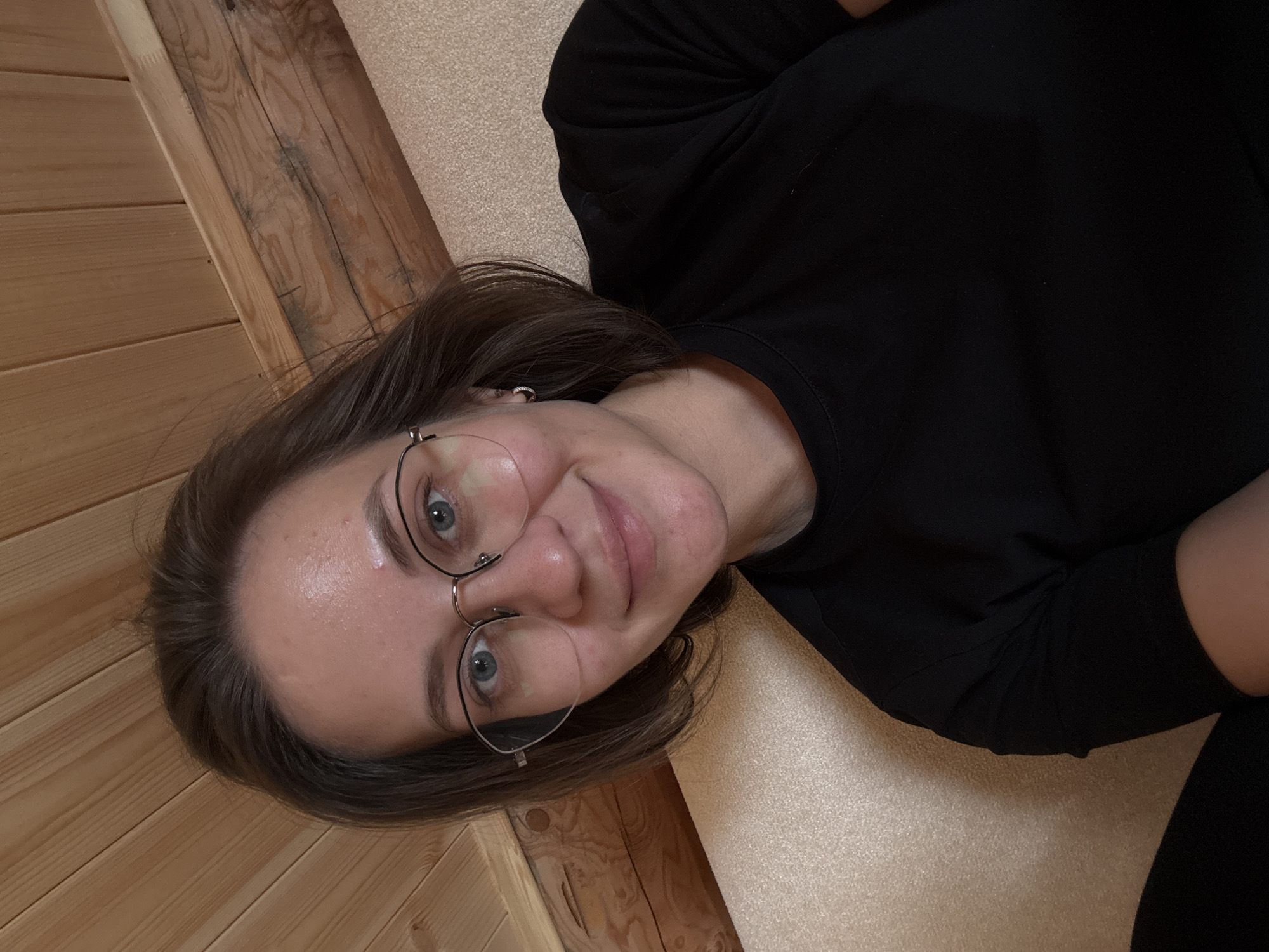 A female individual, appearing to be in her 20s or 30s, is captured in a headshot-style self-portrait, smiling gently towards the camera. She has shoulder-length brown hair, parted to her left. She wears a pair of wire-rimmed glasses with clear lenses and a small silver-colored hoop earring in her visible left ear. Her attire consists of a plain black short-sleeved top. The background reveals a rustic interior setting in the city of Lenino, Russia. The upper portion consists of light-toned wooden paneling, characteristic of wall or ceiling cladding. The lower portion features a textured, light beige or cream-colored upholstered surface, likely a sofa or chair.