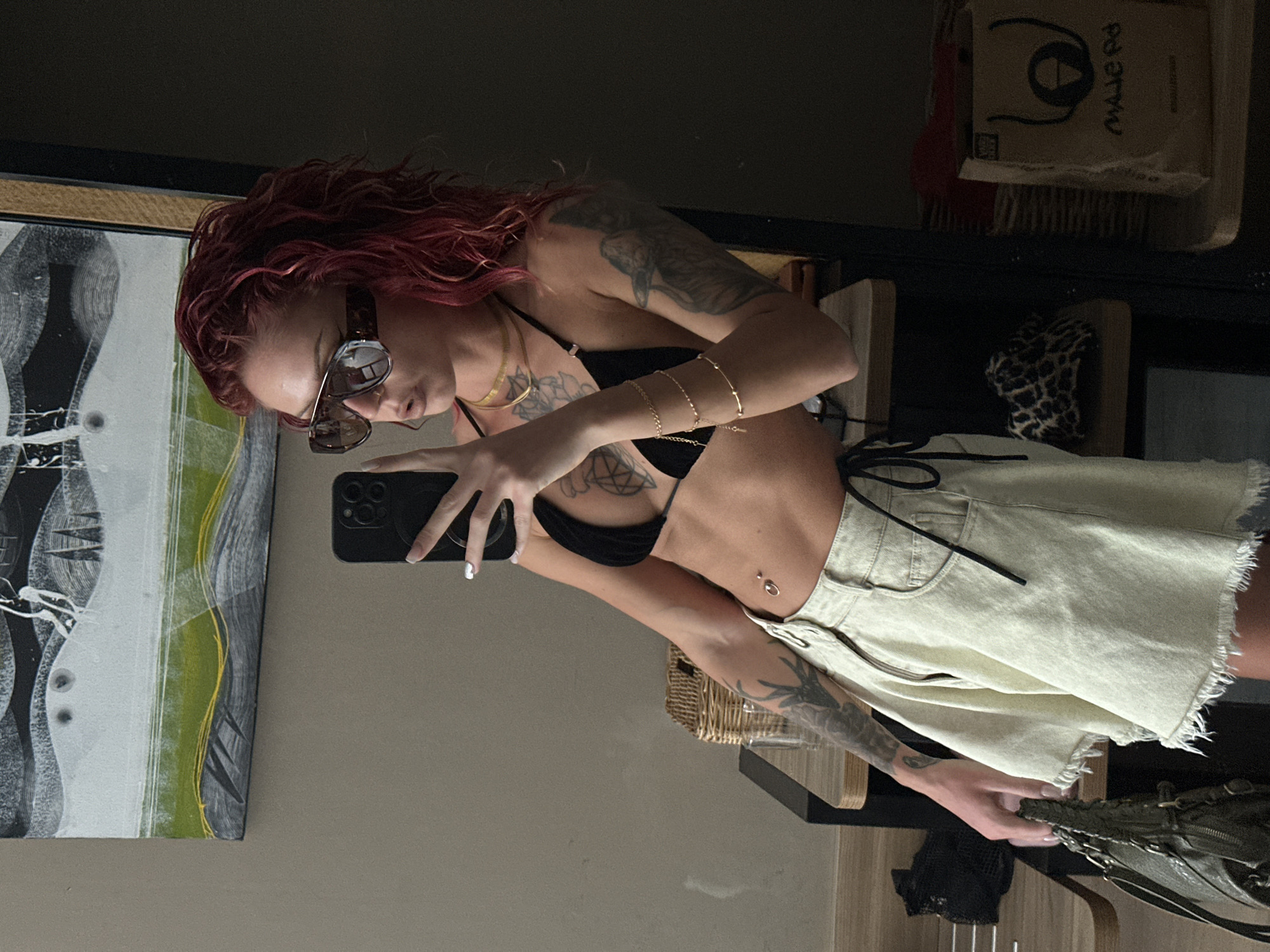A woman with reddish-brown hair is taking a selfie indoors using a black smartphone. She is wearing sunglasses and a black bikini top paired with light-colored denim shorts. The woman has multiple tattoos on her arms and chest, along with gold bracelets on her left wrist and a gold necklace. Behind her is a shelf with miscellaneous items, including boxes and what appears to be animal print fabric. A framed piece of abstract art hangs on the wall to her left. The environment appears to be a room with neutral-toned walls. The city and country are unknown.