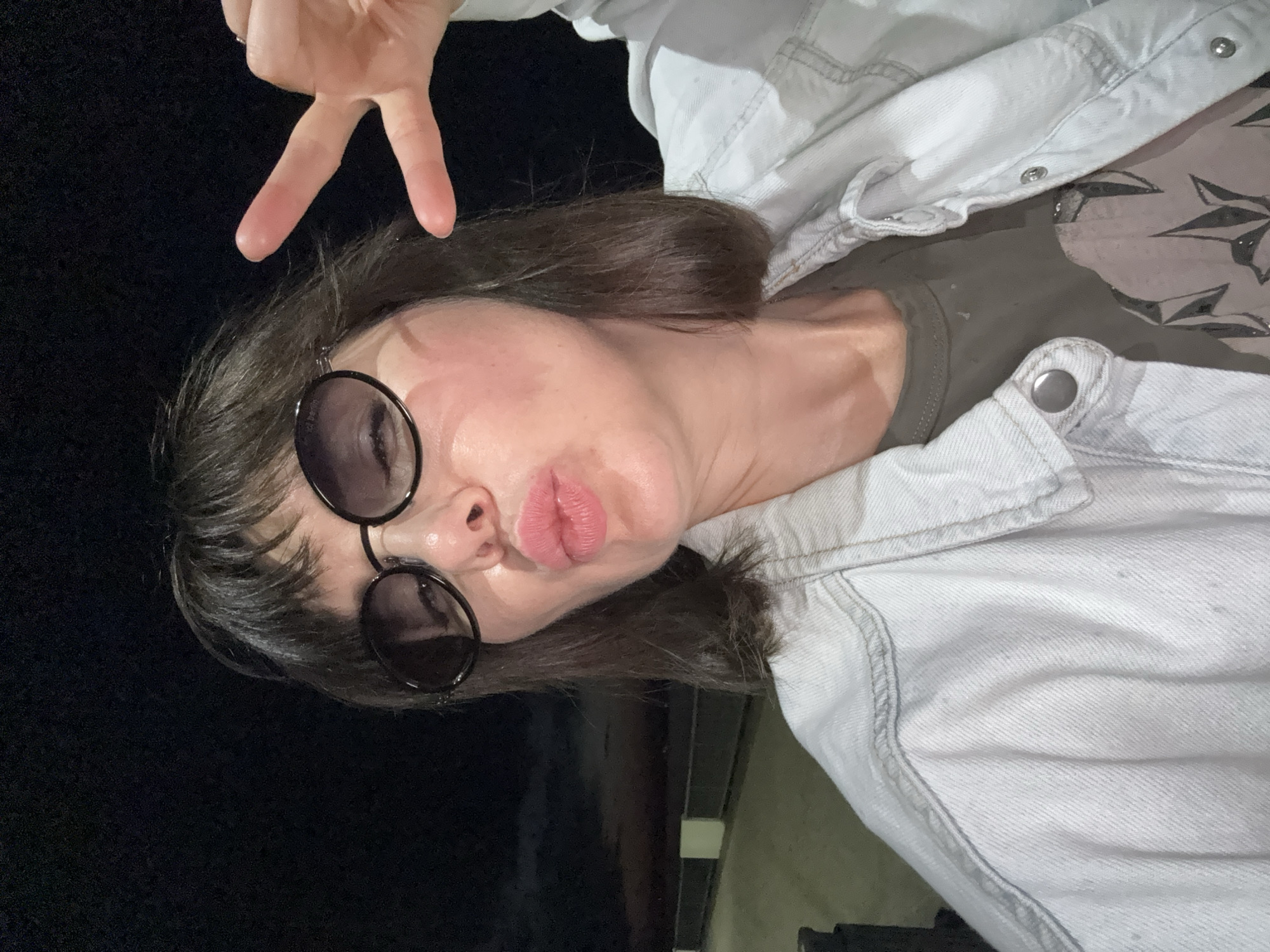 A woman with dark hair and bangs, wearing round sunglasses and a light-colored denim jacket over a dark t-shirt, puckers her lips and holds up two fingers in a peace sign while taking a selfie outdoors at night. The background shows a dark sky with some clouds and a faint horizon line, suggesting an outdoor location. The environment appears to be a public space, possibly a promenade or waterfront, given the presence of a low wall or railing in the background.