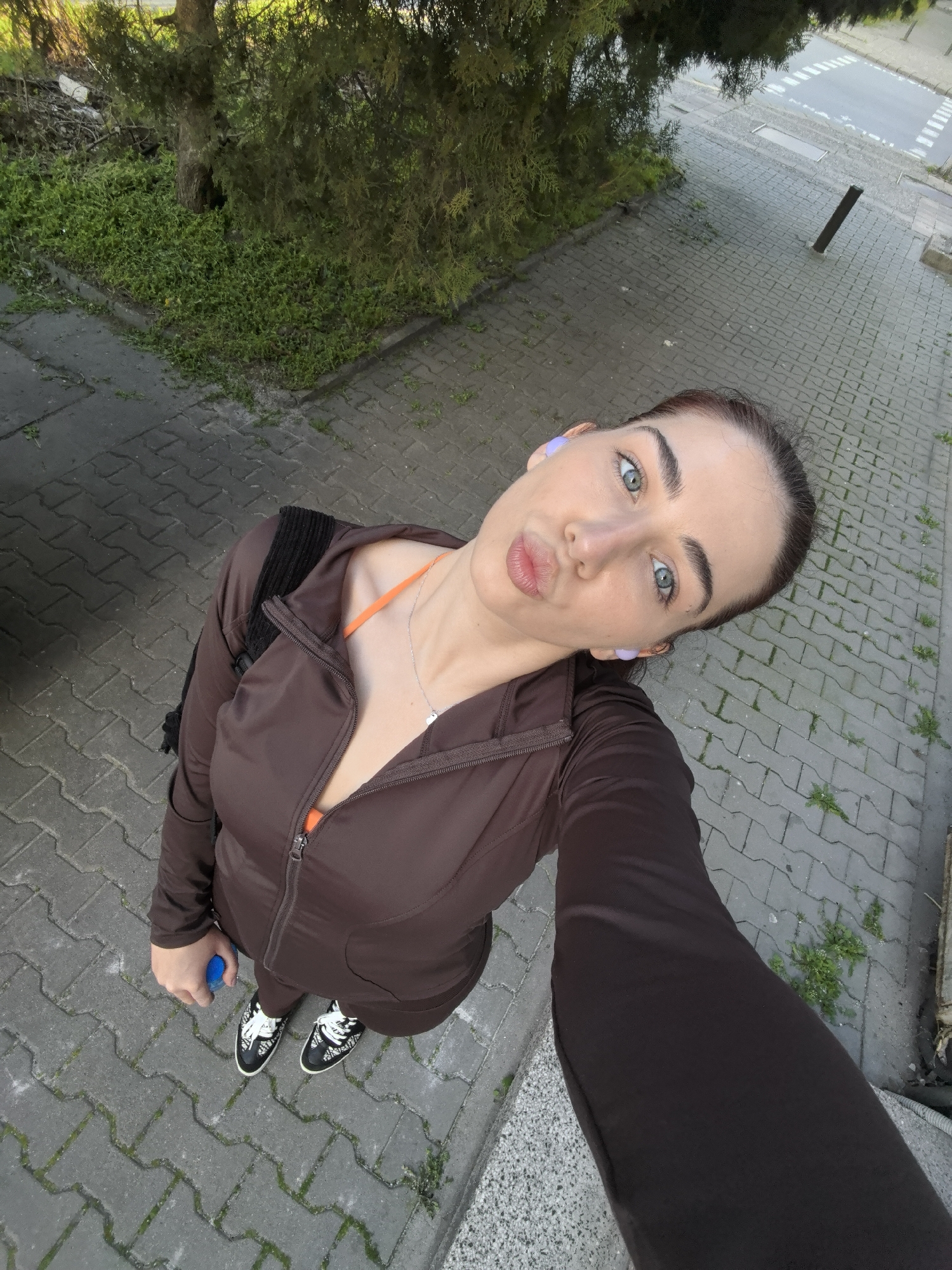 A woman stands on a gray paved street with greenery along its edges. She is wearing a brown zip-up jacket, an orange top visible at the neckline, dark pants, and black-and-white sneakers. She has a backpack on her back. She is holding a blue water bottle in her left hand. She appears to be taking a selfie, with her head tilted and lips puckered. The setting appears to be a street in an urban environment, possibly a park or residential area. The specific location within Stara Zagora, Bulgaria is not discernible from this image alone.