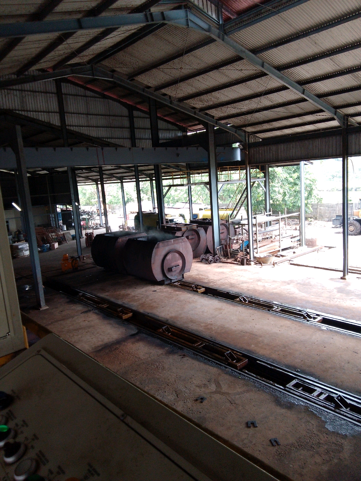 The image shows the interior of a large industrial workshop or factory, likely located in Gunung Manaon, Indonesia. The structure has a corrugated metal roof supported by steel beams and columns, with open sides allowing natural light and ventilation. The floor is concrete, showing signs of wear and some debris.
In the foreground, to the left, is a control panel with several buttons and switches, suggesting an automated or controlled process is taking place. In the center of the image, two large, cylindrical metal objects are visible. These appear to be furnaces or large tanks, with one emitting a plume of steam or smoke. They are positioned on a track system, indicating they might be moved or processed.
To the left of the large metal objects, there are various materials and equipment, including stacked metal rods or pipes, what appear to be smaller metal drums or containers, and a visible orange electric motor. In the background, through the open sides of the workshop, lush green trees and foliage are visible, suggesting the location is surrounded by nature. Further in the background, some vehicles, including what looks like a yellow forklift or heavy machinery, can be seen.
The overall scene depicts an active industrial environment engaged in some form of metal processing or manufacturing. The lighting is a mix of natural light coming from the open sides and artificial light from within the workshop.