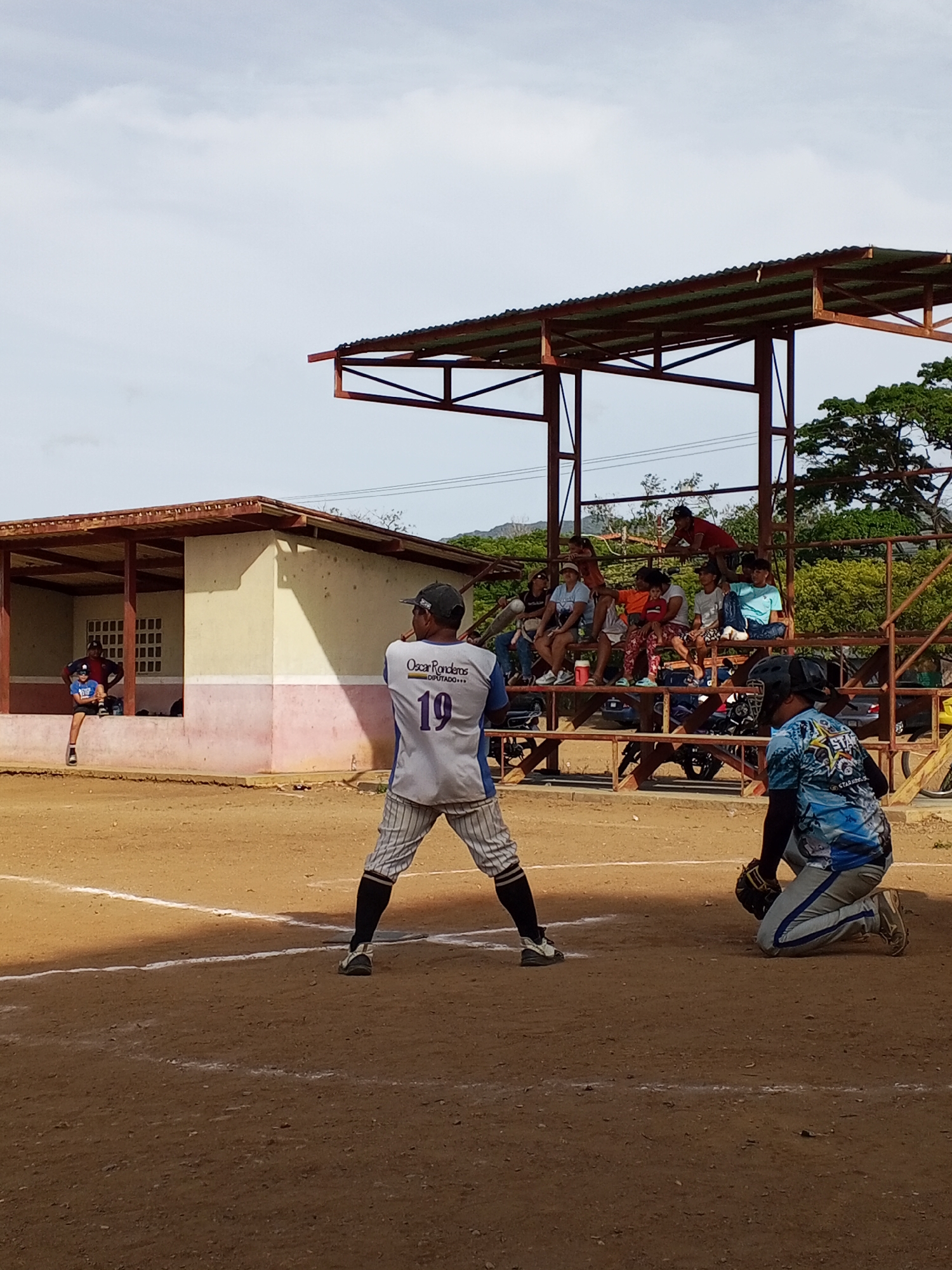 Here is a detailed description of the image:
**Scene:** A baseball game is in progress. The location appears to be a community baseball field in a rural or underdeveloped area. The sky is overcast.
**People:**
* **Batter:** A baseball batter wearing a grey uniform with the number "19" on the back, pinstripe pants, black socks and a cap is in a batting stance.
* **Catcher:** A catcher wearing catcher's gear is kneeling in the catcher's box behind home plate.
* **Spectators:** Several spectators are seated in bleachers behind the catcher.
* **Two people are sitting in the shelter.**
**Objects:**
* **Baseball Bat:** Assumed to be in the batter's hands, although not fully visible.
* **Baseball Field:** A dirt baseball field with painted lines is present.
* **Bleachers:** A metal framed bleachers with a rusty metal roof.
* **Shelter:** A one-room shelter is present behind the batter.
**Actions:**
* The batter is preparing to hit.
* The catcher is in position to receive a pitch.
* Spectators are watching the game.
**Environment:**
* The location is in a rural area with trees in the background.
* The field appears to be in a dry, possibly arid environment.
* The structures appear to be old and in need of repair.
**Location:**
* City: Unknown
* Unknown