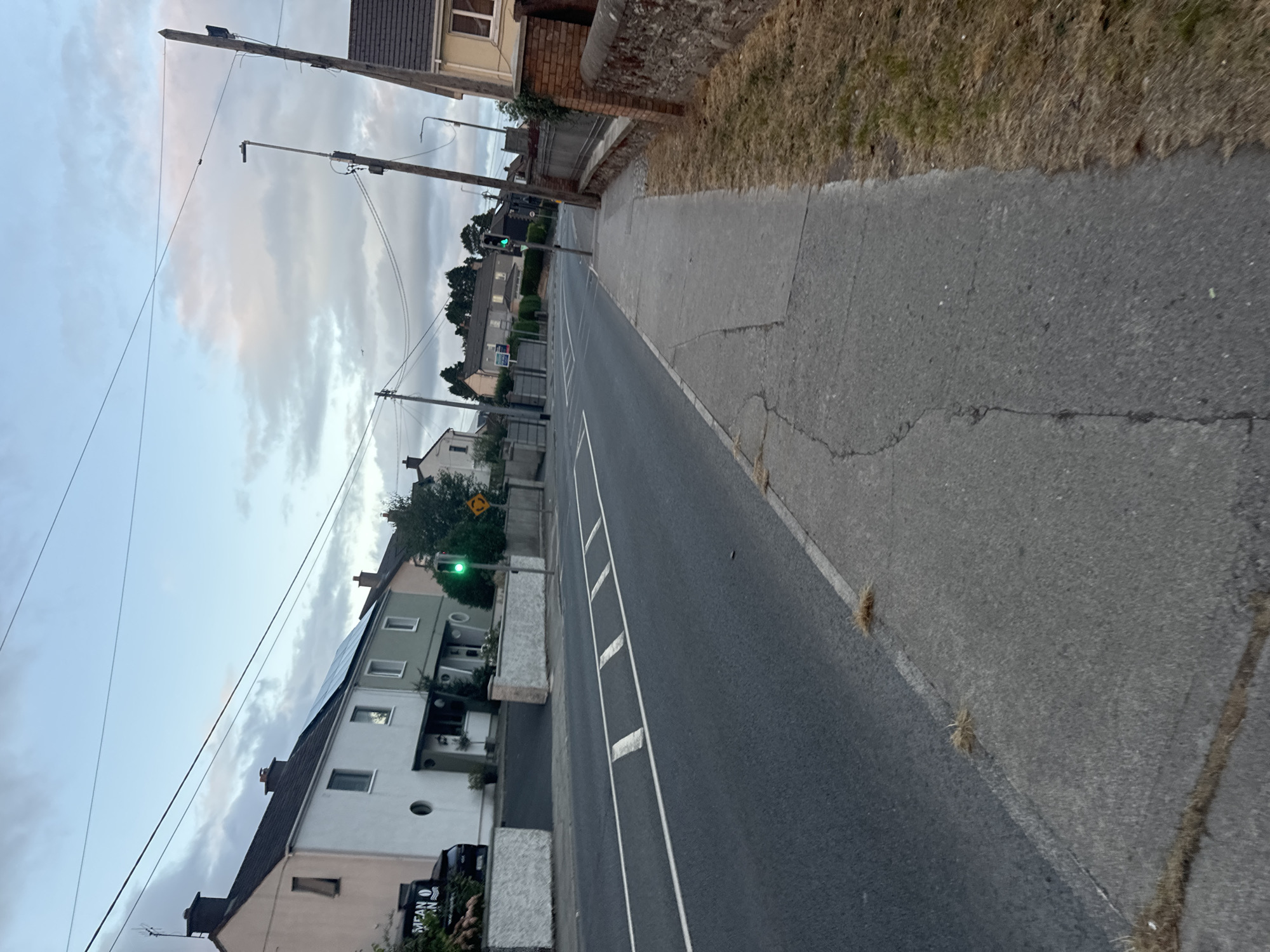 The image shows a street scene in Waterford, Ireland, likely during daytime with overcast conditions. The perspective is from a sidewalk looking across a two-lane road. On the left side of the road, a row of terraced houses with varying colored facades and solar panels on one roof are visible. Some houses have white decorative walls in front of them. A black vehicle is parked in front of one of the houses.
On the right side of the road, a concrete sidewalk is present, bordered by a low brick wall. Beyond the wall, dry grass and some foliage can be seen. There are several utility poles with wires running overhead throughout the scene. A traffic light is visible on the left side of the road, currently displaying a green light. A yellow sign with a circular arrow indicating a roundabout is also present near the traffic light. The road surface appears to be asphalt with white line markings, including what appear to be speed bumps or rumble strips. The sky is cloudy, with some hints of pink and orange suggesting it might be early morning or late afternoon. The overall impression is of a residential street in a town or city.