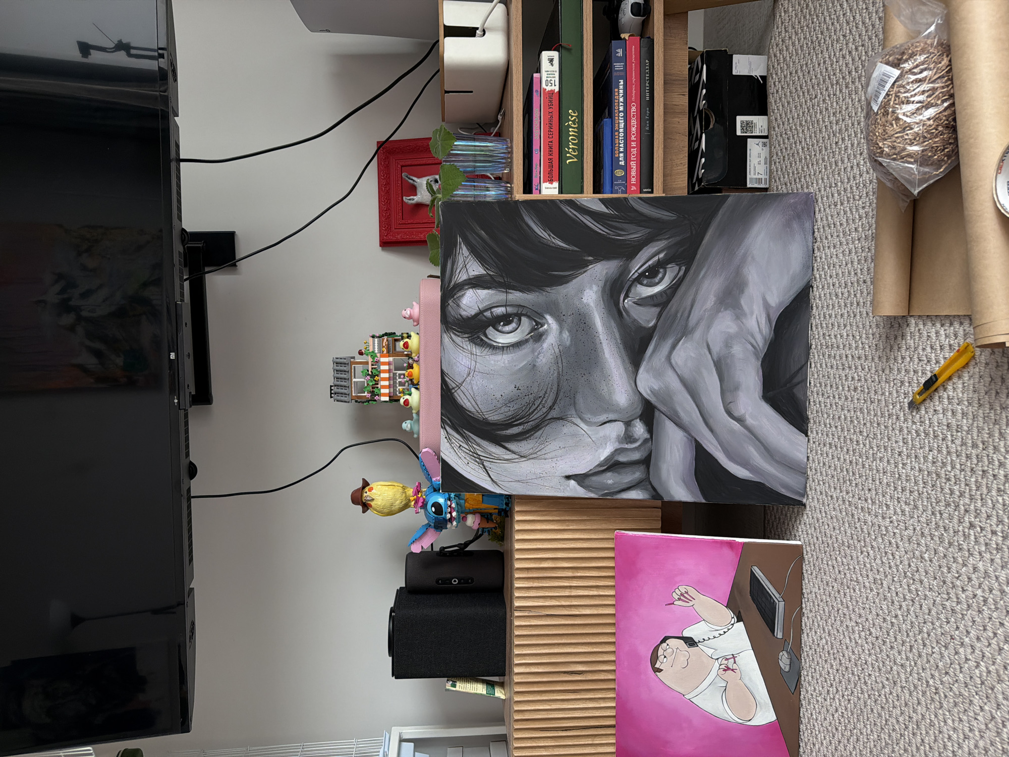 The image depicts an indoor scene, likely a living room, with several art pieces and decorative items. In the foreground, two paintings are visible. The one on the left is a pink canvas featuring a cartoon character resembling Peter Griffin from Family Guy, depicted at a desk with a computer and phone. The painting on the right is a black and white, close-up portrait of a person's eyes and face, rendered in a realistic style.
Behind the paintings, on a wooden media console, are two black speakers, a yellow bird figurine with a hat, a blue Stitch figurine, and a small Lego building. Above the console, a large flat-screen television is mounted on the wall.
To the right of the console is a wooden shelving unit. On the shelves are several books, including one with the title "Véronese" and another in Russian, along with two glasses containing plant cuttings and a white cable management box. A red picture frame is also present. Below the shelves, a black shoebox is visible.
The floor is carpeted in a grey, textured pattern. In the bottom right corner, a yellow utility knife and rolls of brown paper are present, suggesting ongoing art or craft activities, possibly related to framing or packaging the artwork. The overall impression is that of a personal space, perhaps an artist's studio or a creative living area. The image does not contain any specific visual cues that would indicate a location within Krasnoyarsk, Russia.