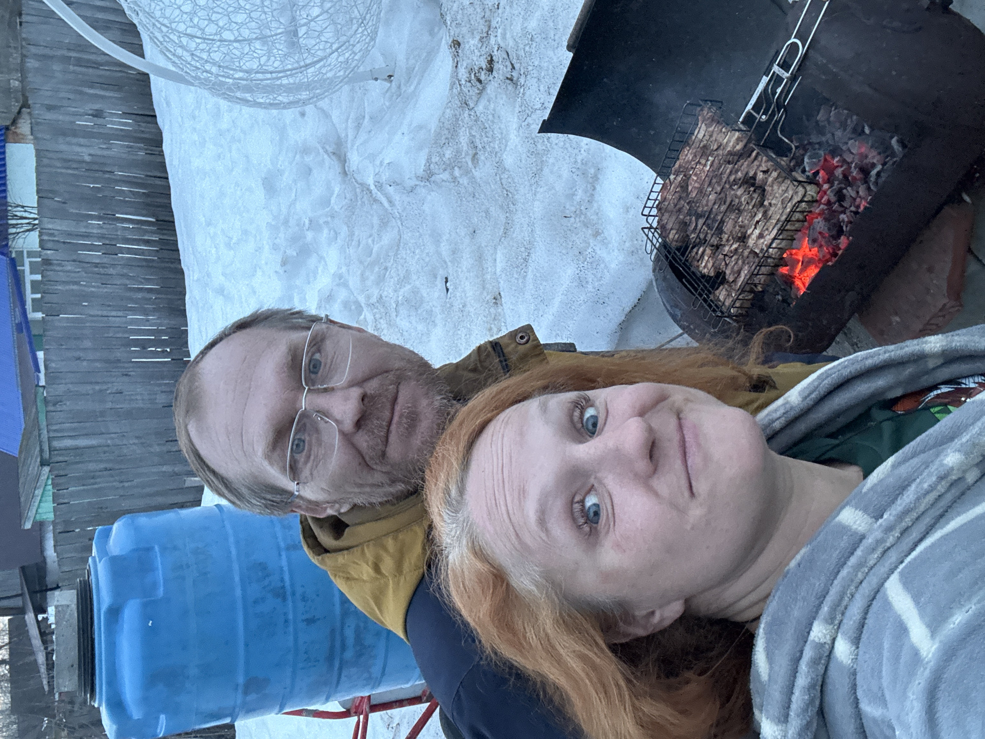In Chaikovsky, Russia, two individuals are captured in a selfie-style photograph outdoors during what appears to be late winter or early spring. A woman with reddish-orange hair and visible gray roots occupies the foreground, looking directly at the camera with wide blue eyes and a slight smile. She wears a grey and white patterned plush robe over a green garment. Behind her, a man with a beard and mustache, wearing glasses and a mustard-yellow jacket over a dark blue garment, smiles faintly towards the camera.
To their right, a dark, cylindrical barbecue grill, seemingly fashioned from a cut barrel, contains glowing red coals and a metal rack laden with multiple pieces of cooking meat. The grill rests on a reddish support object, possibly a brick. The ground around them is covered in uneven patches of snow. In the background, to the left, a large, blue plastic water tank on a red frame is partially visible. To the right, a white, intricately woven or wire-frame spherical object, possibly a chair or decorative element, is partially visible against the snow. A weathered wooden fence runs across the mid-background, separating the immediate area from distant residential-style buildings with various roof colors and sparse vegetation. The overall lighting suggests dusk or an overcast sky.