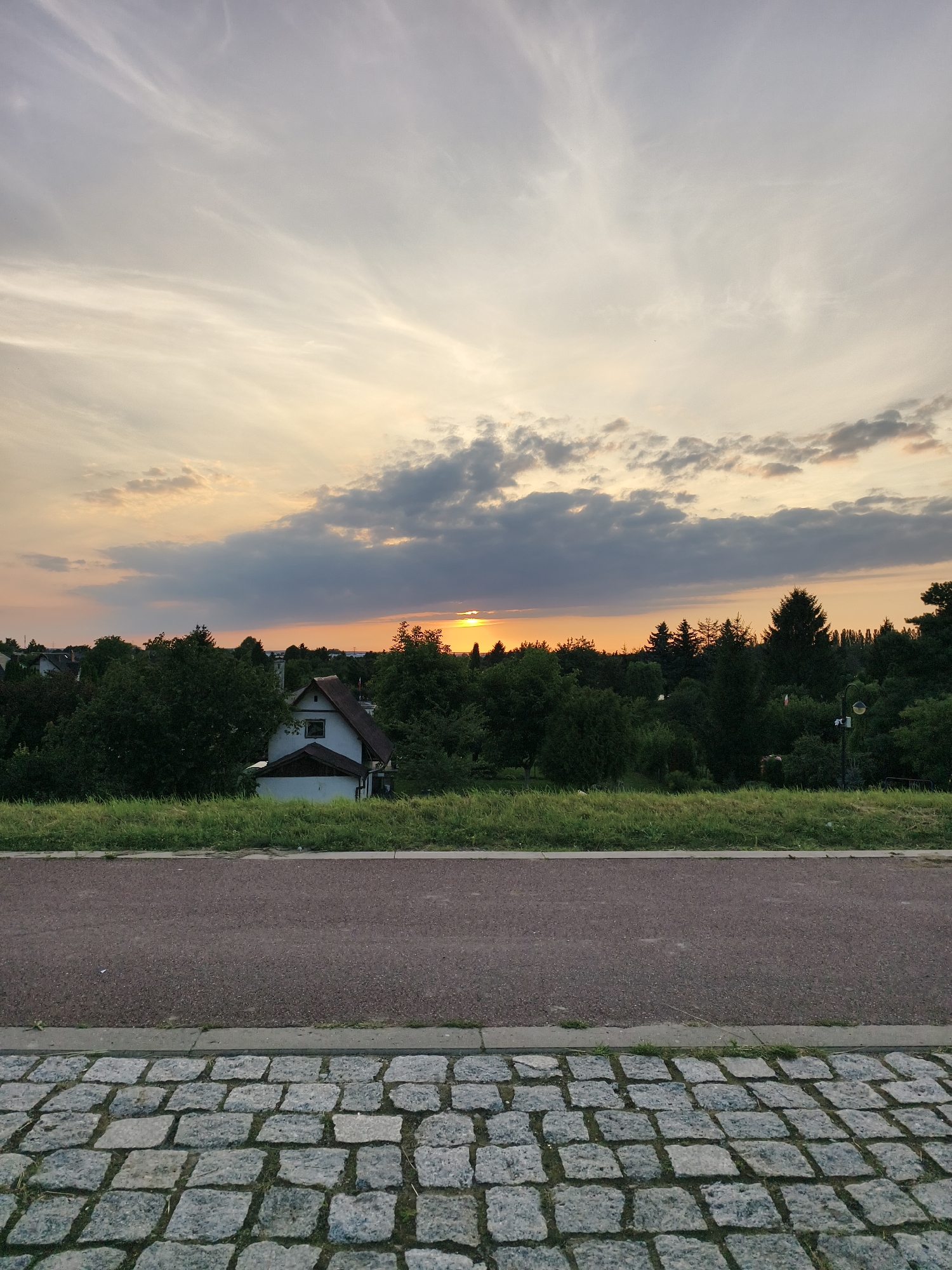 A landscape photograph captures a sunset over a densely wooded area with a few houses peeking through the trees. The sky is a mix of pale orange and gray clouds, with the sun a bright orange disc near the horizon. In the foreground, a paved path runs horizontally across the image, bordered by a strip of green grass. The path is made of dark asphalt, and beyond it, a section of cobblestone pavement is visible, creating a textured foreground. A solitary, light-colored house with a steeply pitched roof is partially visible among the trees, to the left of the center of the image. The overall scene suggests a peaceful evening in a suburban or rural setting. The location is Tczew, Poland.