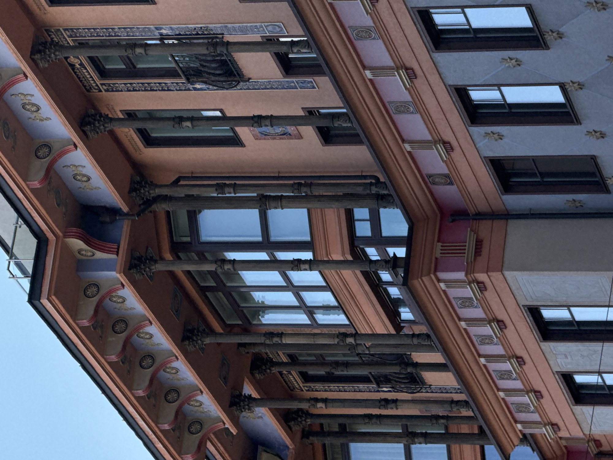 The image captures an upward-angled view of a building in Moscow, Russia, showcasing a portion of its exterior architecture. The building features ornate details, including decorative columns, painted ceilings with circular motifs, and window patterns. The architectural style appears to be of a classical or neo-classical design, with distinct ornamentation and symmetry. The building's facade is primarily pink and blue, with decorative elements highlighted in gold. The presence of windows with visible reflections indicates the time of day, likely daytime, with clear weather.
