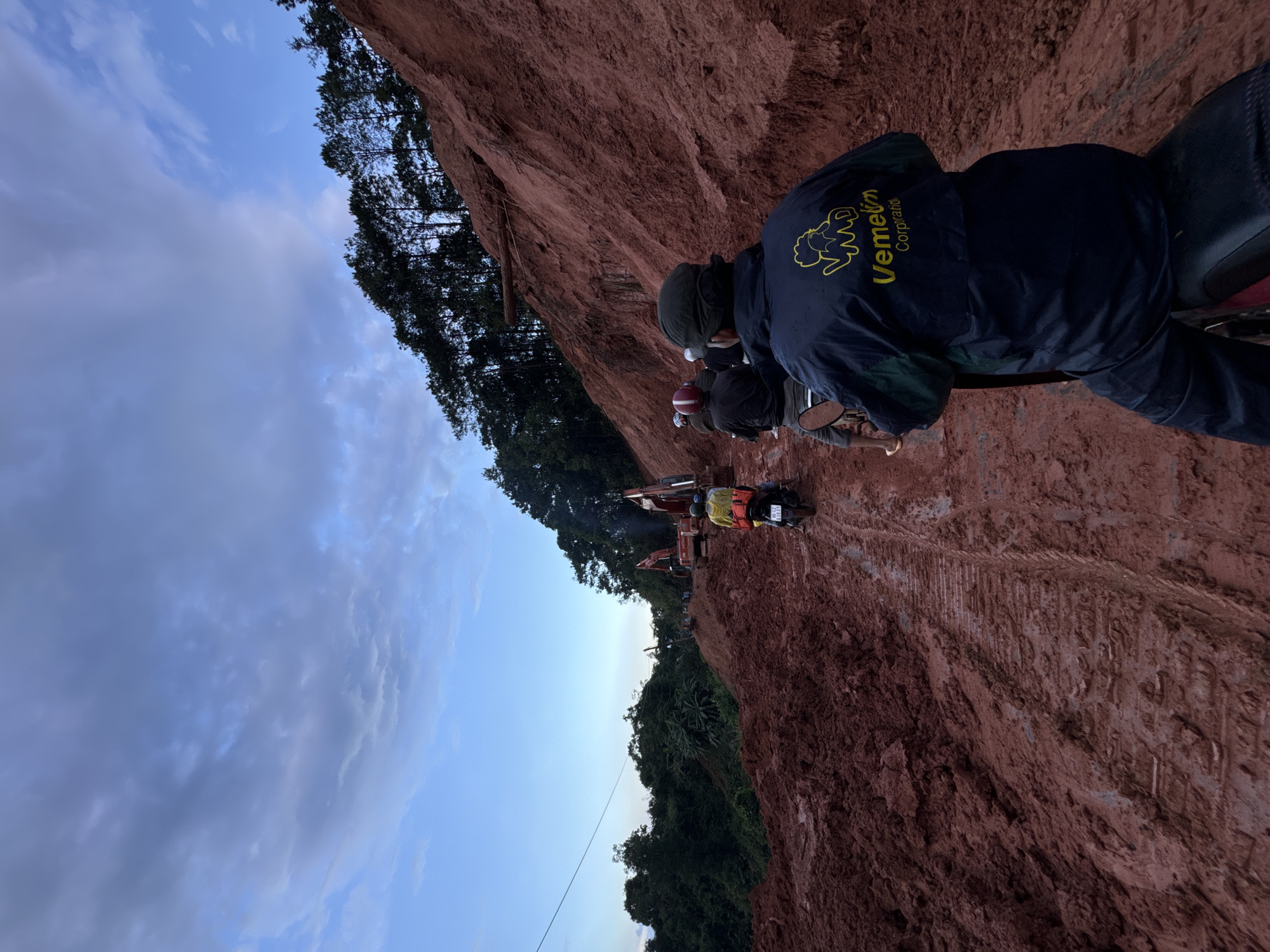 In Nao Sẻ, Vietnam, a dirt road carved through a recently excavated hillside is occupied by several motorcycles and construction equipment. The ground is a deep reddish-brown mud, heavily rutted by the tracks of heavy machinery, indicating ongoing construction or landslide remediation. The sky above is a gradient of blues and grays, with scattered clouds suggesting it is either early morning or late evening.
In the foreground, a person is riding a motorcycle, facing away from the camera. They are wearing a dark blue jacket with a yellow logo that reads "Vemelin Corporation." Behind this rider, two other motorcycles are visible, carrying passengers. The second motorcycle has a rider wearing a red helmet and a passenger in a yellow jacket with a red backpack. The third motorcycle has a rider wearing a white cap.
Further down the muddy track, two large excavators are actively working. One is a red Hitachi excavator, and another appears to be a similar construction machine. Trees line the top of the hill to the left and right of the road. The overall scene depicts a challenging and active construction site in a rural Vietnamese setting.
