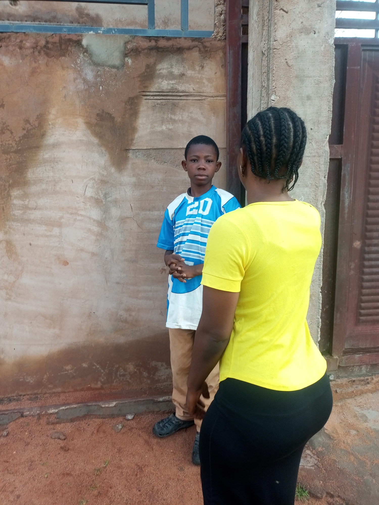 A young Black male stands in front of a textured, light brown wall with some staining and discoloration. He is wearing a blue and white striped t-shirt with the number "20" on it and tan pants. His hands are clasped in front of him. To the right of the frame, a Black female stands with her back to the camera. She has her hair styled in braids and is wearing a bright yellow t-shirt and black pants. The ground is covered in reddish-brown dirt and pebbles. A concrete pillar and a dark brown wooden door are visible behind the female. The scene appears to be outdoors in a residential or public area.