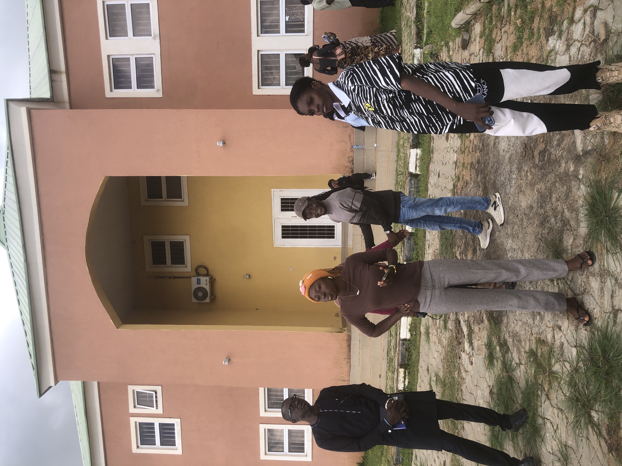 In front of a multi-story building with peach and yellow facades and a green, ribbed roof, a group of individuals stand on a paved area with patches of grass. To the left, a man wearing a black traditional outfit and glasses looks off to the side, holding a blue book. Beside him, a woman in a brown long-sleeved shirt and gray trousers, wearing an orange beanie, holds a smartphone and looks forward. To the right, a person in a black and white striped shirt and black and white color-blocked pants stands facing away from the camera, holding a phone. Other individuals are visible in the background, some walking or standing near the building's entrance. The building features several windows with white frames and metal bars. An air conditioning unit is mounted on the yellow section of the building. The overall scene suggests an outdoor gathering or meeting on a cloudy day. The location is identified as Gwafan, Nigeria.