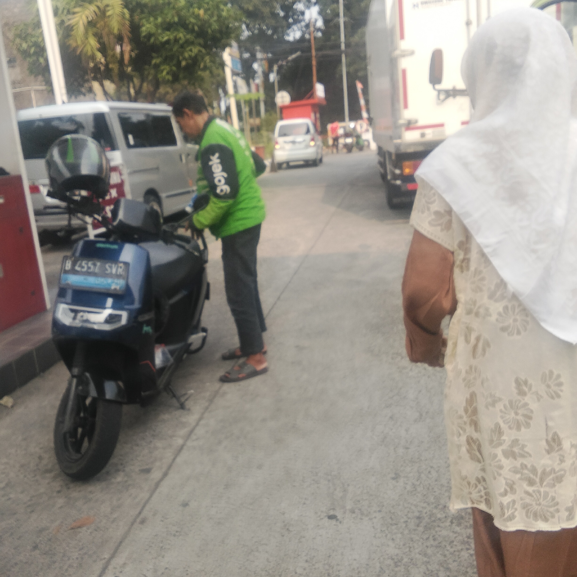 The image captures an outdoor scene in Cibinong, Indonesia, likely at a gas station or a similar service area. A Gojek delivery rider, wearing a green jacket with the company logo on the sleeve and dark pants, is attending to a blue motorcycle. The motorcycle has a license plate that reads "B 4557 SVR". In the background, a white van is parked, and further down the road, other vehicles, including a white car and a truck, are visible. A woman wearing a white headscarf and a light-colored patterned top is walking away from the motorcycle, towards the right side of the frame. The environment appears to be a paved area with some greenery and trees in the background.