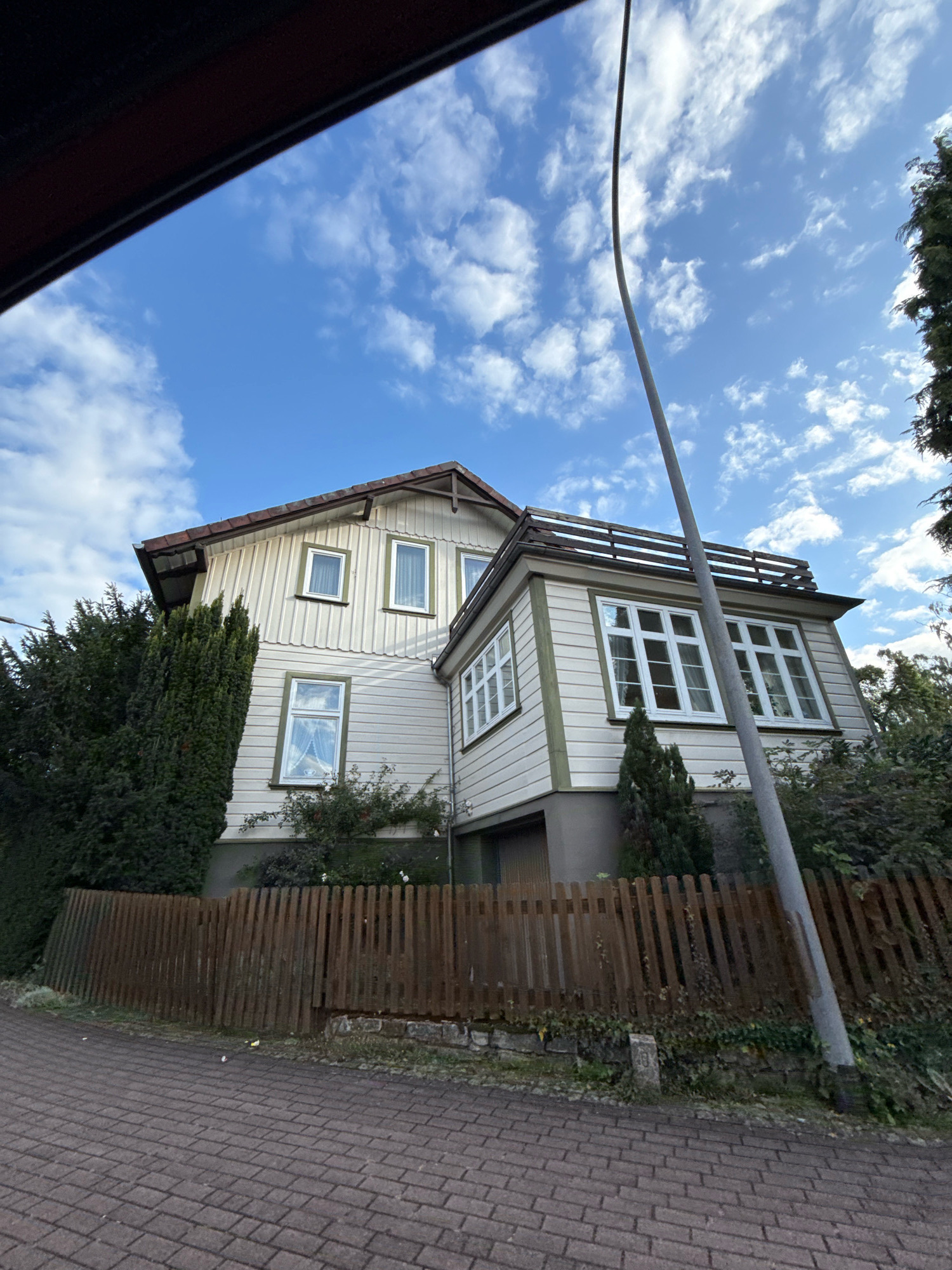 The image shows a two-story house with white siding and green trim, situated in Osterode am Harz, Germany. The house has a gabled roof and multiple windows, some with white curtains visible. A wooden fence runs along the front of the property, with dense green trees and bushes surrounding the house. A grey lamppost is positioned in front of the house, leaning slightly. The ground in front of the fence is paved with brickwork. The sky is blue with scattered white clouds.