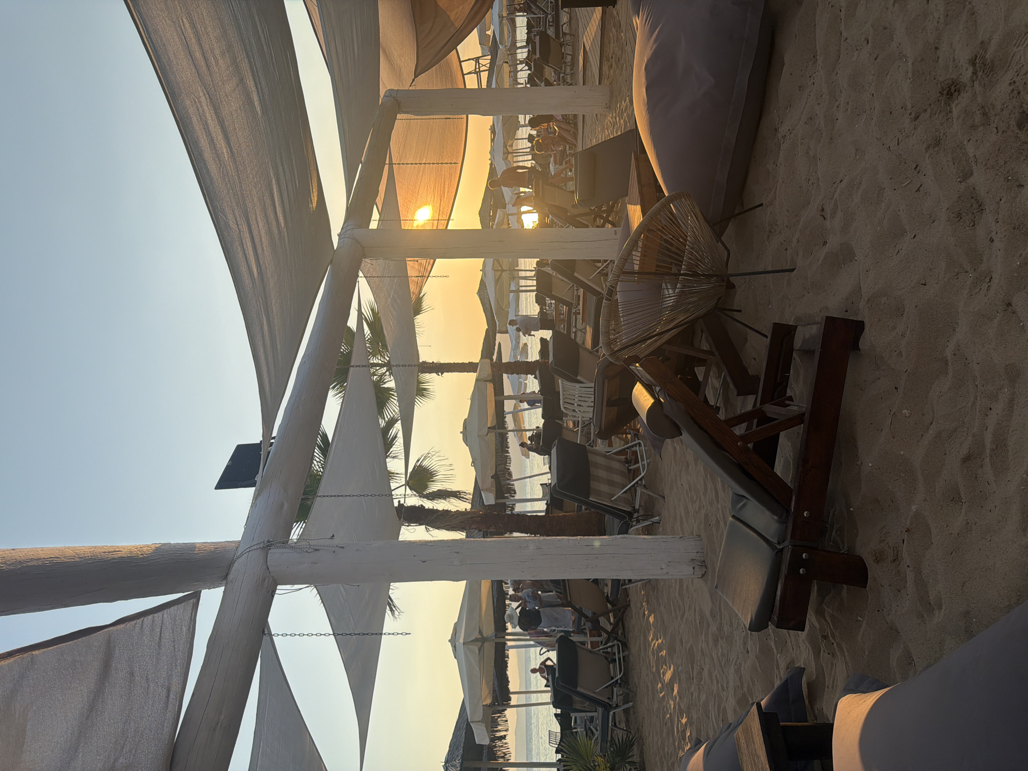 The image depicts a serene beach scene at Ofrynio Beach, Greece, during sunset. The foreground is dominated by sand, with several lounge chairs and large floor cushions arranged for relaxation. A distinctive woven rattan chair is visible on the right. In the mid-ground, rows of sun umbrellas and more lounge chairs are set up, some occupied by people. Beyond the beach, the sea is calm, reflecting the warm hues of the setting sun. Palm trees are scattered throughout the scene, adding to the tropical ambiance. Overhead, fabric canopies are stretched between white wooden posts, providing shade. The sun is low in the sky, casting a golden glow over the entire landscape, and its rays are visible through the gaps in the structures.