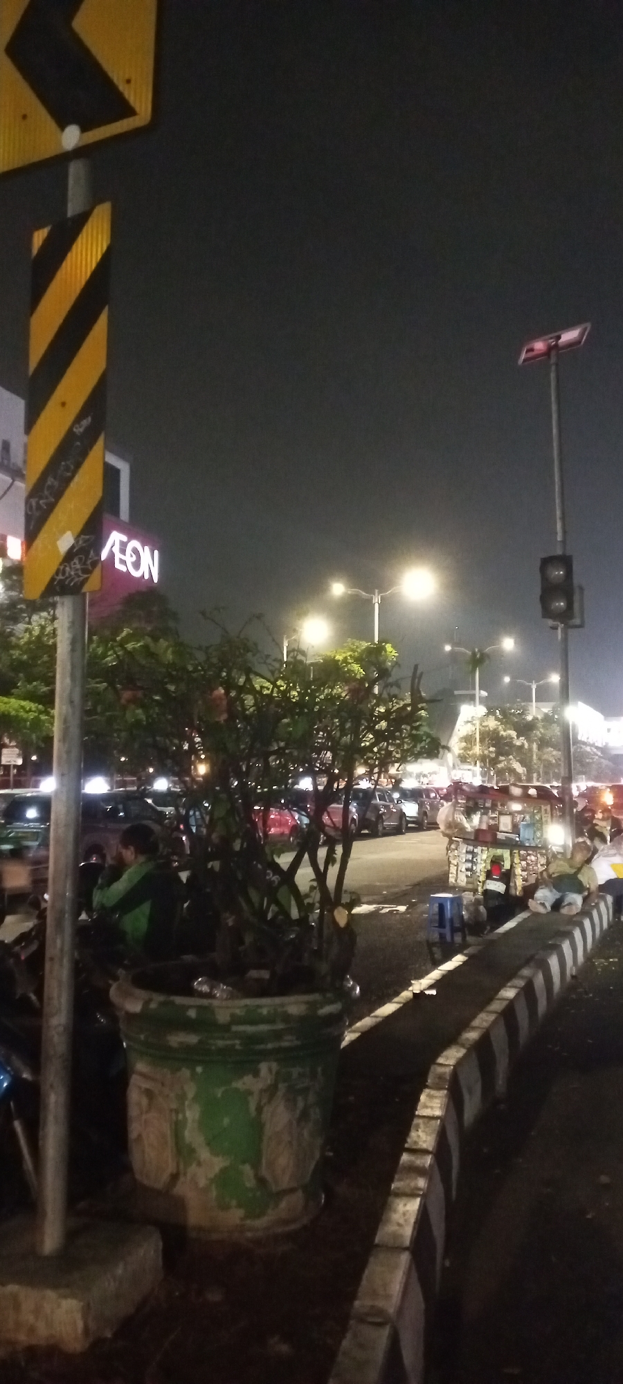 The image depicts an outdoor scene at night in South Jakarta, Indonesia. In the foreground, a large, weathered green planter pot sits on a dark surface. A young person wearing a green jacket is seated on a motorcycle to the left of the planter, facing away from the camera and looking at a tree. The tree itself is in the planter and has some flowering elements. Behind the planter and the person, several cars are parked along a street. To the right of the planter, a sidewalk with black and white striped curbing runs alongside the street. Further down the street, there is a small food cart. In the background, several streetlights illuminate the area, and a traffic light is visible on a pole. A large sign with a yellow and black chevron pattern is present on the left side of the image, attached to a pole. The sign appears to be a directional warning sign. The sky is dark, indicating it is nighttime. The "AEON" logo, illuminated in pink, is visible on a building in the mid-ground, to the left of the street.