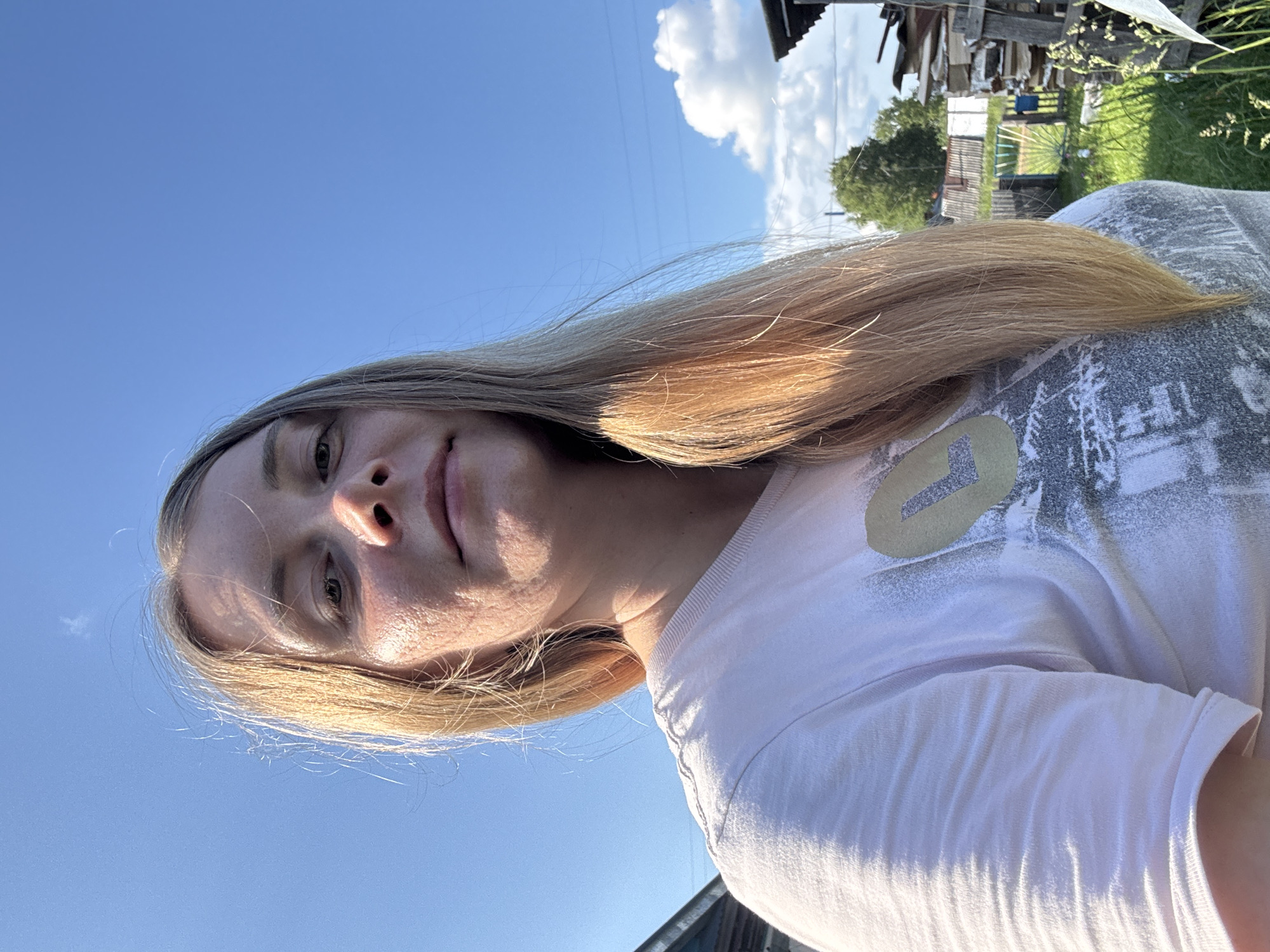 An outdoor, eye-level, close-up, selfie-style shot shows a fair-skinned woman with long, blonde hair and light freckles across her nose and cheeks. She is wearing a light pink t-shirt with a distressed, light gray graphic and a circular emblem with a yellow "L" on the left side of her chest. She is outdoors in front of a bright blue sky with a few wispy white clouds. In the background, several structures are visible, including a wooden building to the left and stacks of lumber and firewood to the right. Green grass and trees are also present. The overall setting appears to be rural, possibly a dacha or countryside home, consistent with a location in or near Kurgan, Russia.