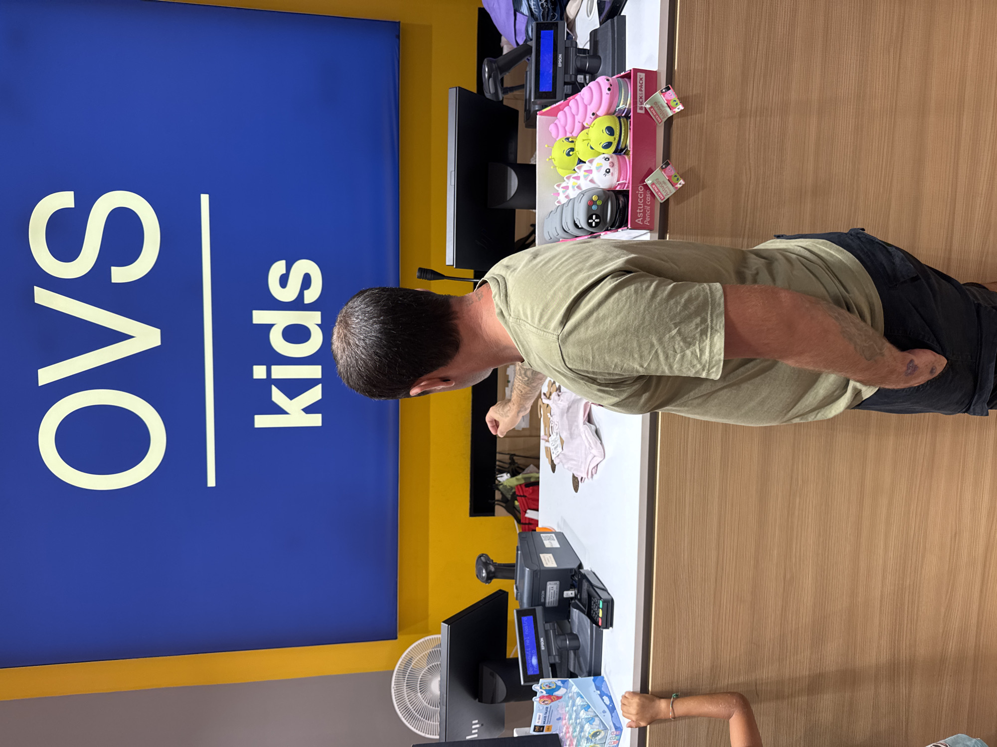 In Carini, Italy, at a retail store featuring a prominent "OVS kids" sign, a man wearing a khaki t-shirt and dark jeans stands at a checkout counter. He is looking down at the counter, with his right hand extended towards a display of novelty pencil cases. To his left, a young child with a bracelet on their left wrist rests their hand on the counter. The counter is equipped with a cash register, a credit card reader, a scanner, and various retail items, including children's clothing and the aforementioned pencil cases. A computer monitor and a fan are also visible behind the counter. The store environment is bright, with a blue and yellow color scheme on the wall signage.