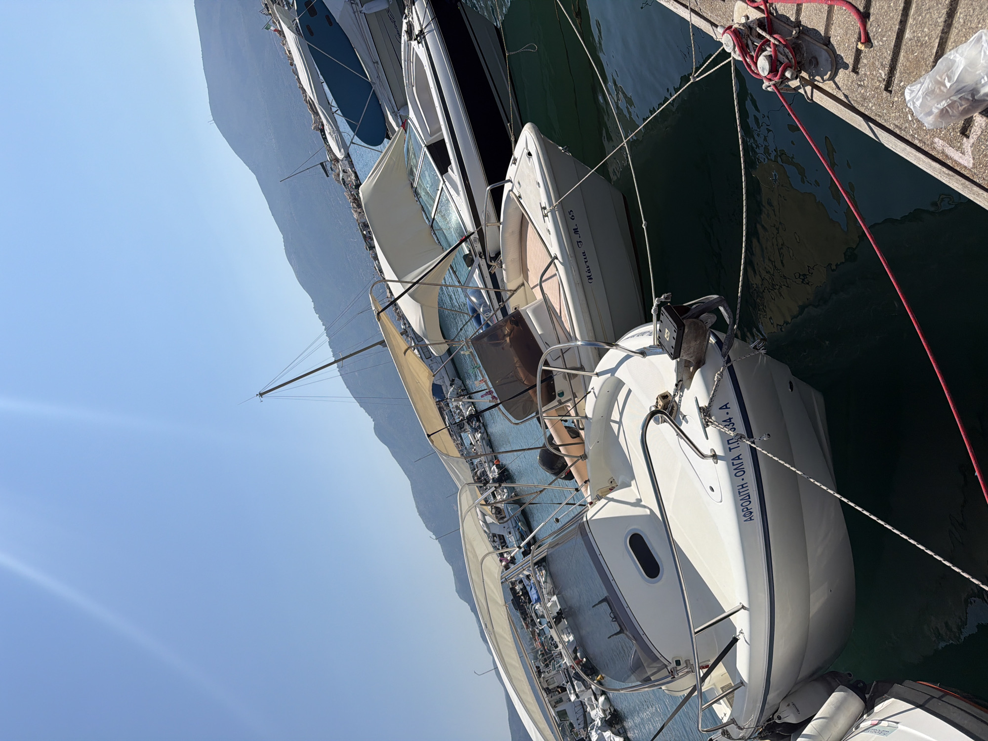 In the Municipal Unit of Alyzia, Greece, a bright, sunny day illuminates a marina filled with several boats docked on calm, dark blue water. The image is dominated by two white motorboats in the foreground, both facing towards the viewer but angled to the right. The boat on the left has a blue stripe along its hull with Greek lettering "ΑΦΡΟΔΙΤΗ-ΟΛΓΑ T.O.334-A". It features a white canopy providing shade over the seating area. The boat on the right is slightly further back and to the right, also with a white canopy, and visible lettering on its hull reads "Μάγισσα T.M. 65". Behind these, numerous other boats are moored, some with canopies and others more visibly covered. In the background, a gentle, hazy mountain range rises against a clear blue sky, with sunlight casting bright flares across the upper portion of the image. The boats are secured to a concrete dock on the right side of the frame, which has a red rope tied to a cleat and a plastic bag lying on its surface.