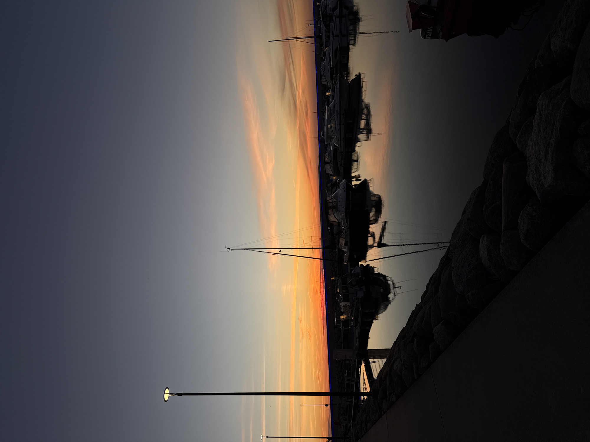 A serene sunset scene unfolds over a marina in Tallinn, Estonia. The sky transitions from a deep, dusky blue at the top to a vibrant band of orange and pink near the horizon, with wispy clouds adding texture. The calm water of the marina beautifully reflects the colors of the sky and the silhouettes of several boats docked along the waterfront.
Prominently featured are various vessels, including sailboats with tall masts and smaller motorboats, their forms softened by the twilight. A concrete walkway lined with large, rough-hewn stones forms the foreground, leading the viewer's eye towards the water. A tall lamppost with a lit globe stands on the left side of the walkway, casting a faint glow. The overall atmosphere is one of tranquility and quiet beauty as day transitions into night. There are no people visible in the frame, suggesting a peaceful and unpopulated moment. The location appears to be a well-maintained harbor or yacht club.