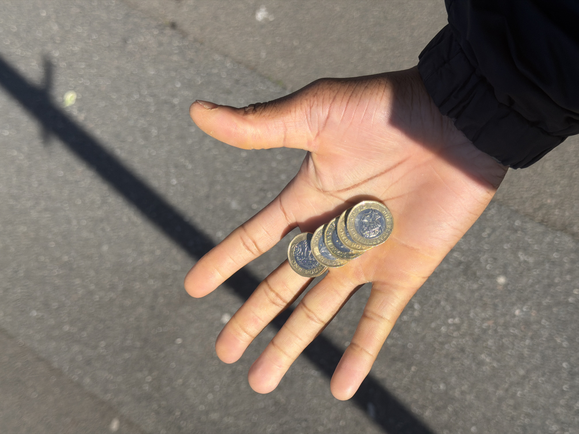 A hand with medium brown skin is holding four British pound coins on its fingers. The coins are stacked on top of the index finger, appearing to be balanced. The person is wearing a black jacket. The background is gray asphalt and the lighting suggests it is outdoors in daylight. A shadow of an object is cast on the asphalt. There is no definitive evidence in the picture to ascertain if the picture was taken in Folkestone or Hythe District in the United Kingdom.