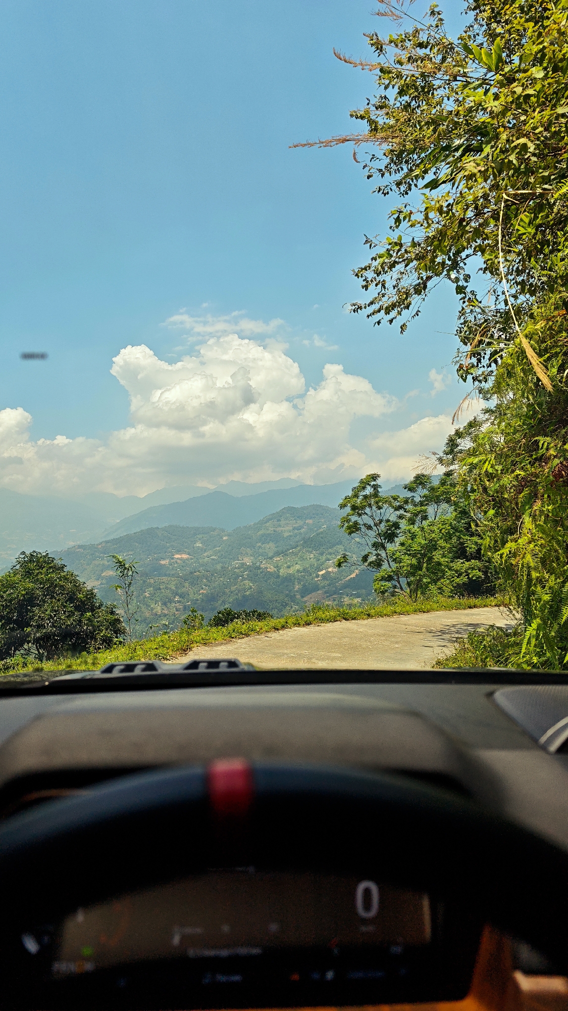 The image shows a view from inside a car driving on a road in Vinh Quang, Vietnam. The car's steering wheel and dashboard are visible in the foreground, displaying a speed of 0 km/h. The road ahead curves slightly to the right and is surrounded by lush green vegetation and trees. In the distance, layers of mountains are visible under a partly cloudy blue sky. The landscape is dotted with settlements and agricultural land.
