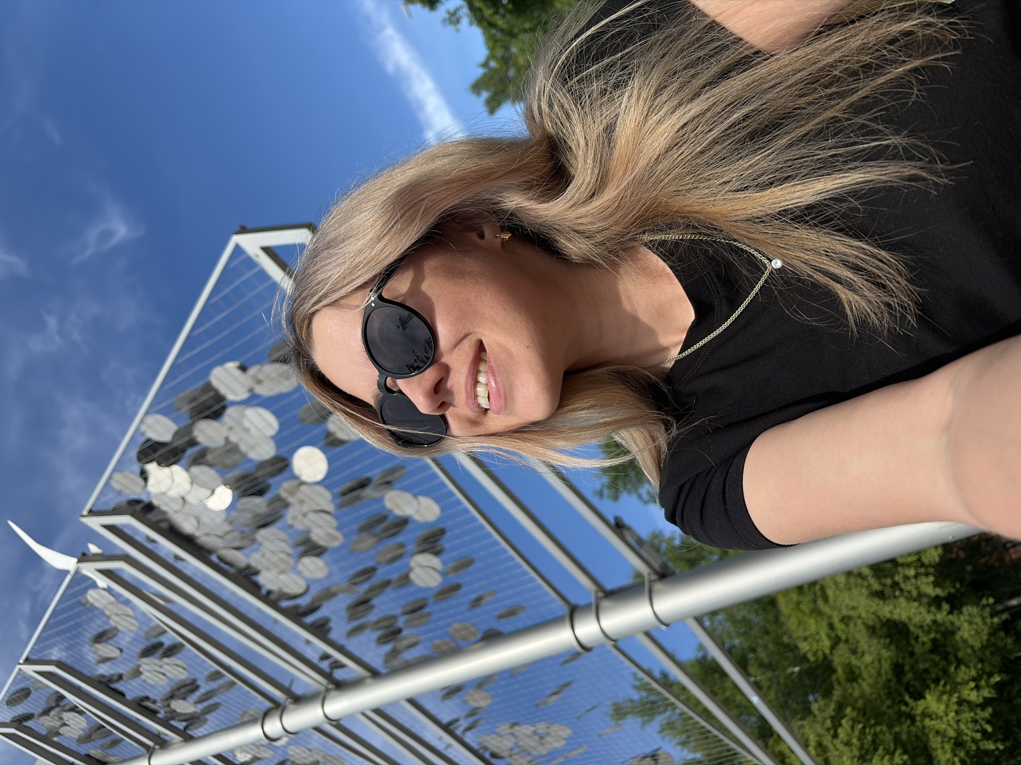 A blonde woman takes a selfie outdoors on a sunny day, smiling broadly into the camera. She wears dark sunglasses, a black top, and a delicate silver necklace with a single clear stone. Her long blonde hair is visible around her face. The image is taken from a low angle, looking upwards towards a clear blue sky.
Behind her, a distinctive metal art installation, known as the "Wishing Tree" or "Tree of Coins" in Chaikovsky, Russia, is prominently featured. This structure consists of multiple tiered rectangular metal frames, within which numerous circular, coin-like metallic objects are attached to a grid. These objects vary in reflectivity and shade, creating a shimmering effect against the blue sky.
The bright sunlight suggests it is midday or afternoon, contributing to the cheerful atmosphere. Green tree foliage is visible in the background, indicating an outdoor, possibly park-like setting. The woman's smile conveys a happy and relaxed mood. No visible text is present in the image.