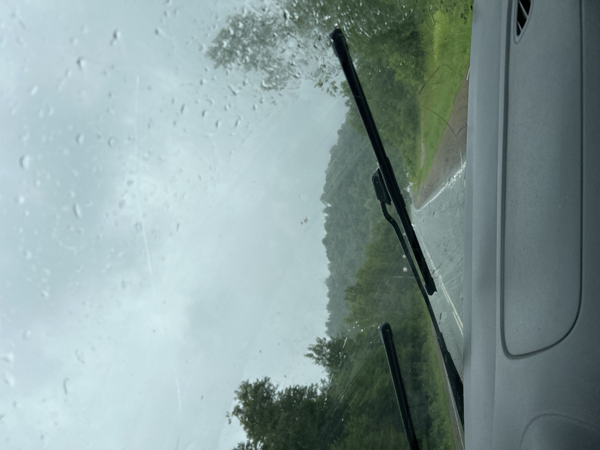 The image shows a view from inside a vehicle looking out through a rain-streaked windshield. Two windshield wipers are visible, one on the left and one on the right, partially clearing the view. The road ahead is wet and appears to curve to the right, surrounded by lush green trees and rolling hills that are shrouded in a misty, overcast sky. The overall impression is one of a rainy day drive through a rural, forested landscape. There is no indication of the city of Кулмас, Russia in the image.