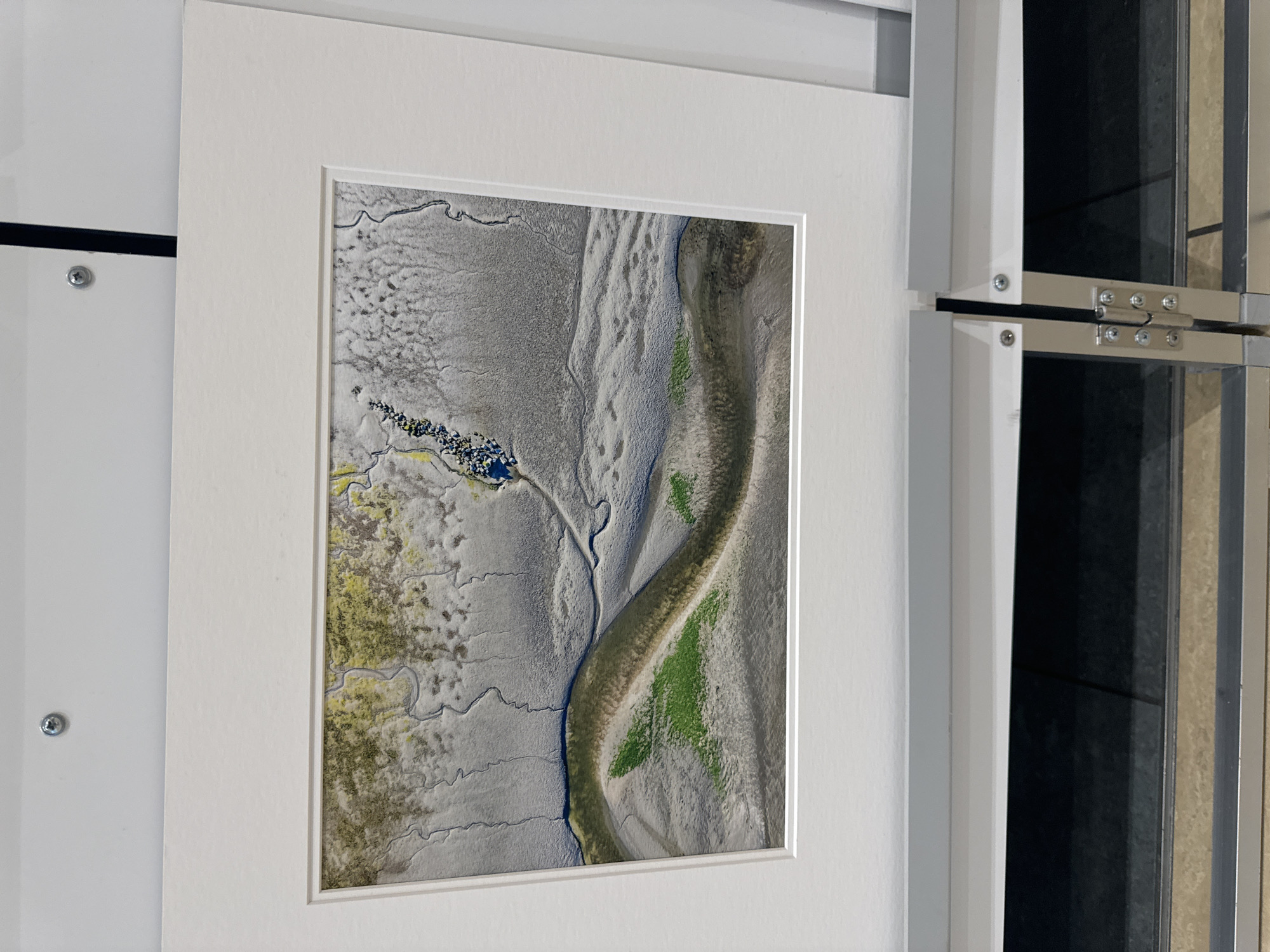 The image displays a framed aerial photograph of a tidal estuary, possibly at low tide, showcasing intricate patterns of mudflats and winding water channels. The photograph is mounted on a white mat and framed by a white border. Patches of green vegetation are visible on some of the mudflats, suggesting areas of marsh or salt-tolerant plant life. In the upper portion of the photograph, small clusters of dark blue and yellow elements suggest either water or debris. The photograph is hung on a plain white wall, with a silver screw visible in the upper left corner. The bottom of the image shows a part of a shelving unit with metal supports and hinges, indicating the photograph is likely displayed in an indoor setting, perhaps a gallery or an office. There are no people, animals, or identifiable landmarks present in the photograph or its immediate surroundings. The location in the city of Kilkenny, Ireland, is not discernible from the image content.
