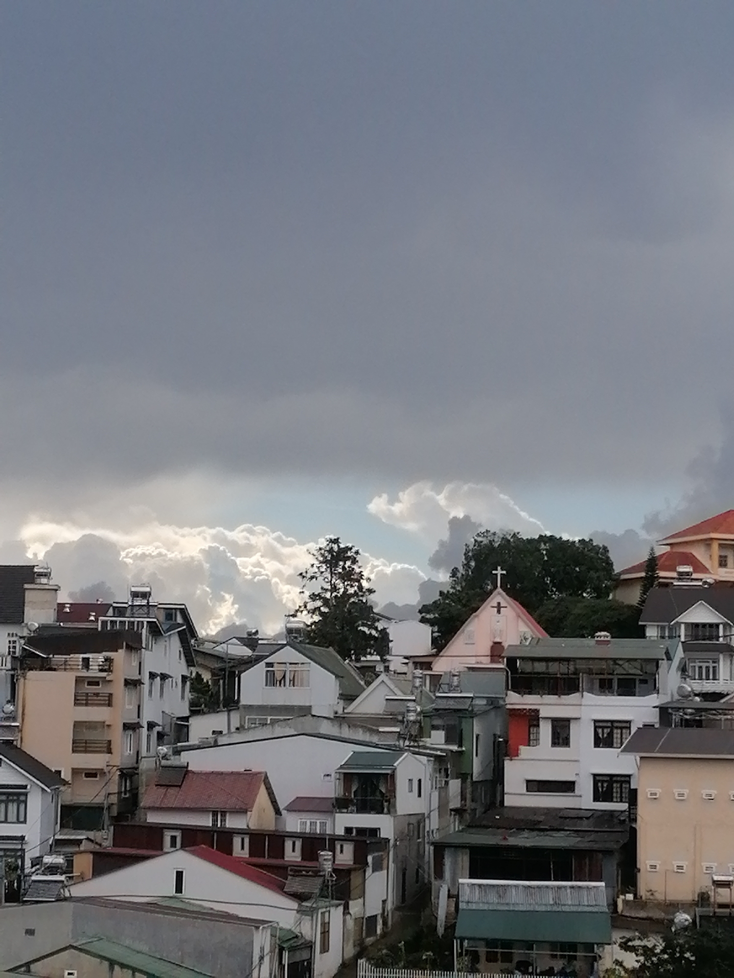 This image depicts a dense urban landscape with a variety of buildings, likely residential, nestled among rolling hills. The architecture suggests a European colonial influence, with multi-story structures featuring pitched roofs, balconies, and various window styles. The scene is dominated by a dramatic sky filled with dark, heavy clouds, hinting at an impending storm. In the foreground, a cluster of buildings exhibits a mix of architectural styles and states of repair. Some are painted in muted colors, while others feature more vibrant hues like orange and red on their roofs. A prominent pink building with a cross at its apex, possibly a church or religious institution, stands out amongst the other structures. The surrounding vegetation is limited, with a few trees visible, one of which is a tall, slender pine. The overall impression is one of a densely populated hillside town, possibly experiencing inclement weather. The presence of structures and architectural elements suggests a location within Lang Biang - Da Lat, Vietnam, a region known for its French colonial architecture and hilly terrain.
