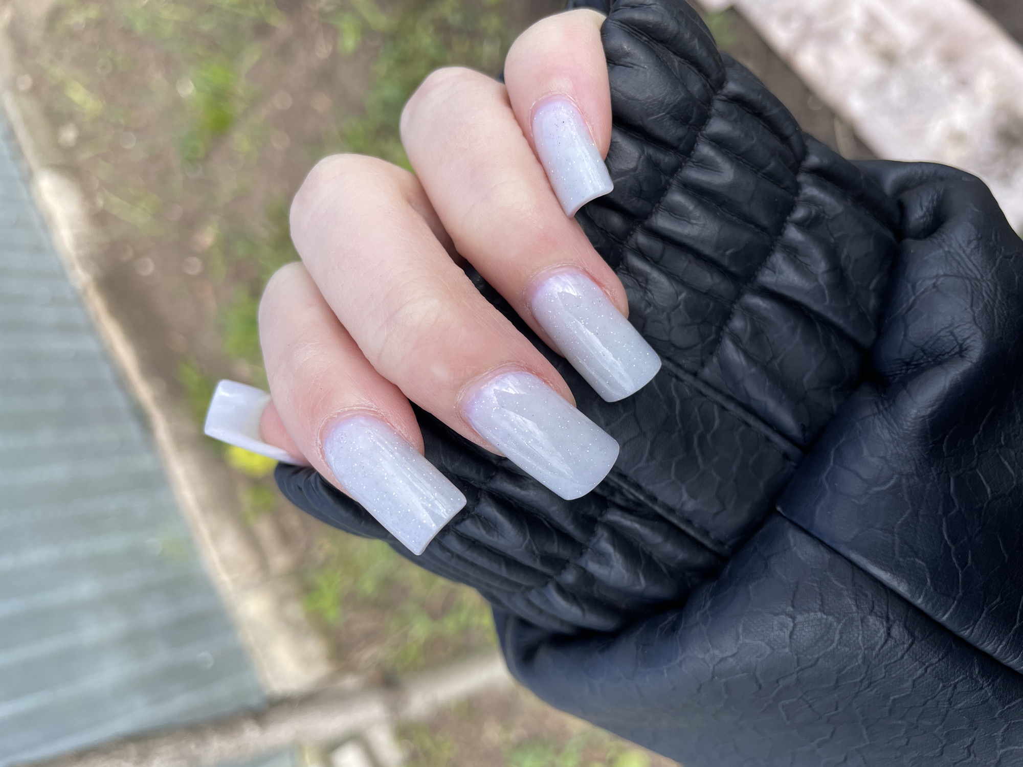 A hand with long, square-shaped acrylic nails painted in a shimmering light grey is resting on a black, quilted leather glove. The hand appears to be of fair-skinned complexion. The background features a blurred outdoor scene, suggesting a location with greenery and pavement. Given the query, this scene is hypothetically situated in Oktyabrsky, Russia.