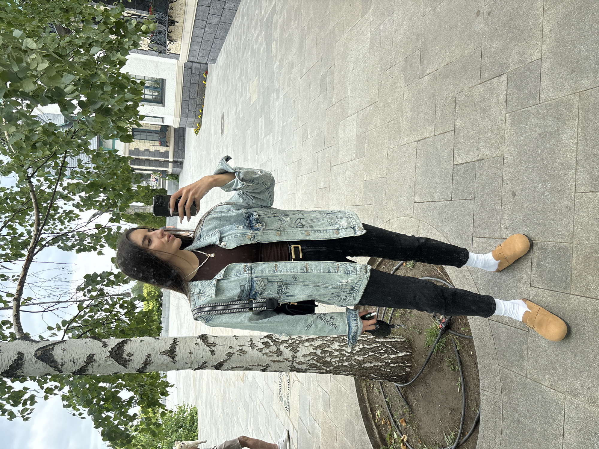 A young woman is captured in a full-body, portrait-style shot, standing outdoors in what appears to be a city square or pedestrian walkway. The image is oriented vertically. She is wearing a light blue denim jacket with intricate graffiti-like designs and dark trousers, paired with tan slip-on shoes and white socks. A dark brown top is visible beneath her open jacket, and she is accessorized with a delicate pearl necklace and a black crossbody bag with a white striped strap.
In her right hand, she holds a black smartphone, suggesting she is either taking a selfie or documenting her surroundings. Her left hand is partially visible, also near the phone. The woman has dark hair and is looking slightly away from the camera, with a thoughtful or relaxed expression.
The setting features a paved ground made of large, grey stone tiles, with a distinct pattern. To the left of the woman, a young birch tree with its characteristic white, patterned bark is prominent, its green leaves providing a natural element. In the background, there is a building with light-colored walls, dark windows, and some architectural detailing. The overall ambiance suggests a pleasant, possibly daytime scene, with soft, diffused lighting. The presence of trees and the paved ground indicate an urban or semi-urban outdoor environment. The image provides no explicit clues about the time of day or weather beyond the general brightness.
No text is clearly visible or legible within the image. The location context is Khryashchevka, Russia.