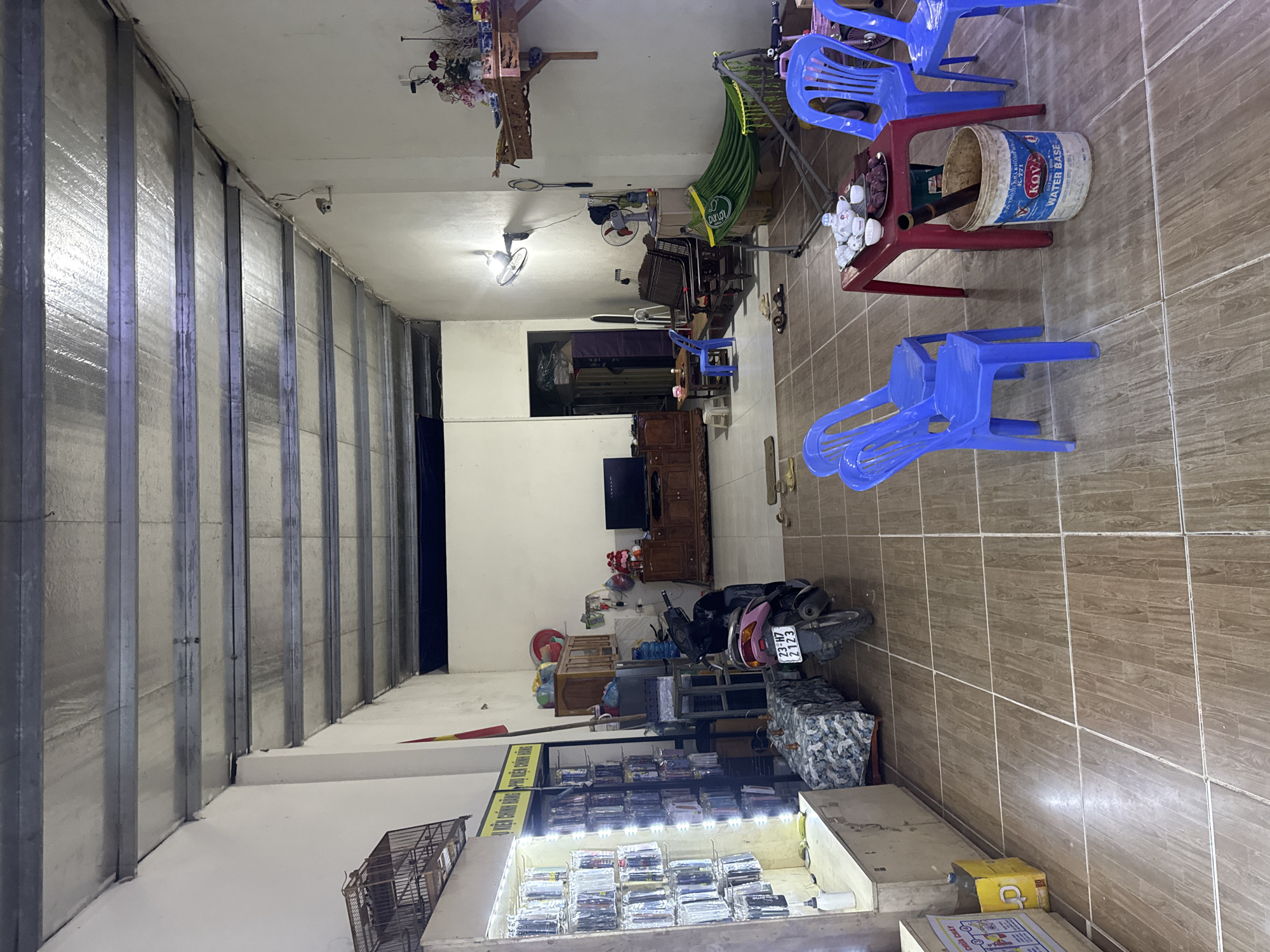 The image depicts the interior of a commercial space in Du Già Commune, Vietnam. The room is cluttered with a variety of items. Along the left side is a corrugated metal wall. The space contains an assortment of objects including blue plastic chairs, a red table with teacups and a paint bucket on it, a hammock, a television, and a motorcycle. The front of the space contains retail shelving. The floor is tiled. A cage and other items are at the front of the store.