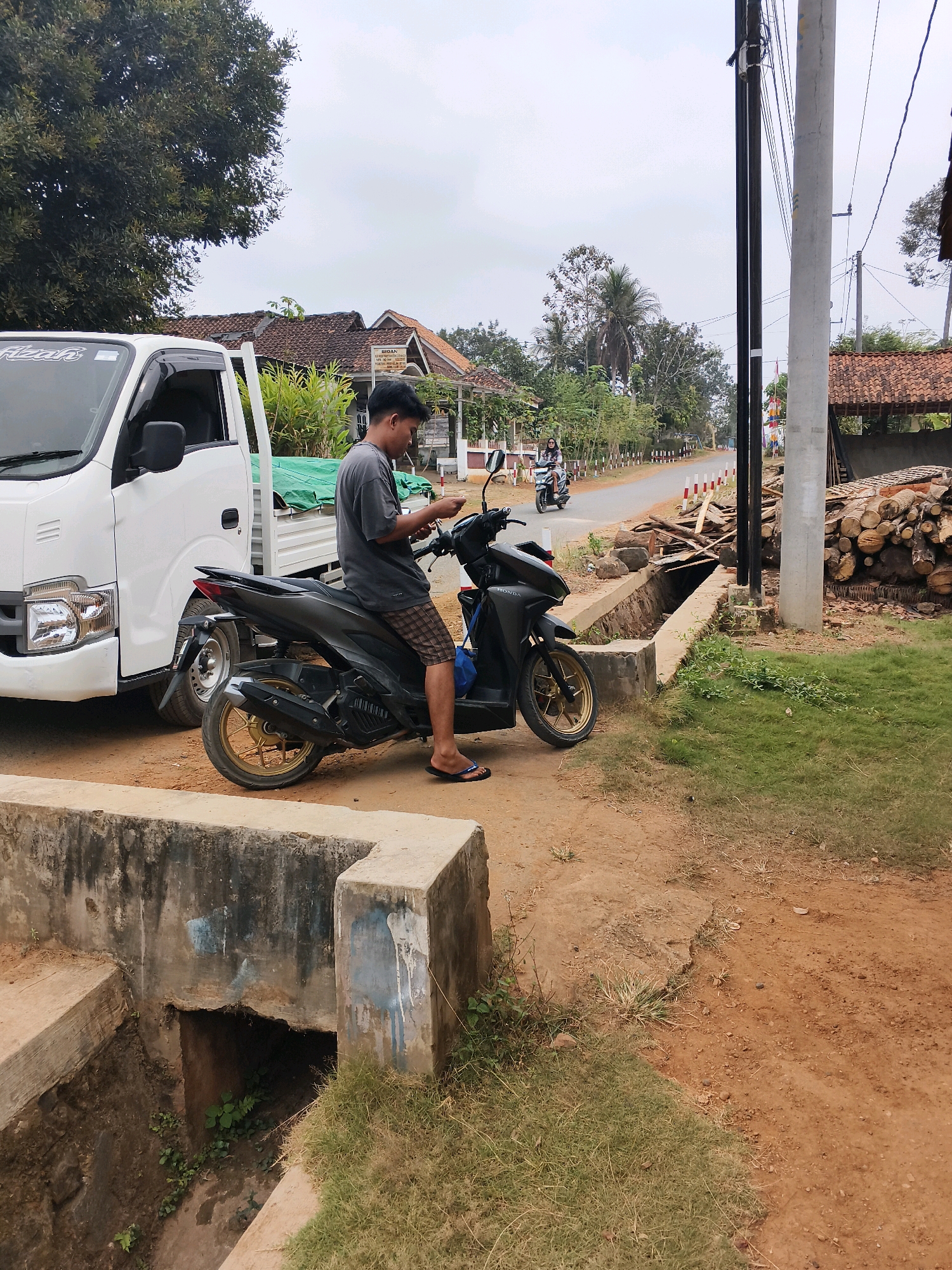 A young man in a gray t-shirt and checkered shorts is seated on a black scooter, looking at his phone. He is positioned on a dirt road next to a white truck. The scooter has gold rims. In the background, a road is visible with other houses and trees. To the right, a stack of logs is piled high next to a utility pole. The scene appears to be a rural or semi-rural area in Banyuwangi, Indonesia, possibly on the outskirts of the city or in a village. A concrete drainage ditch is in the foreground.
