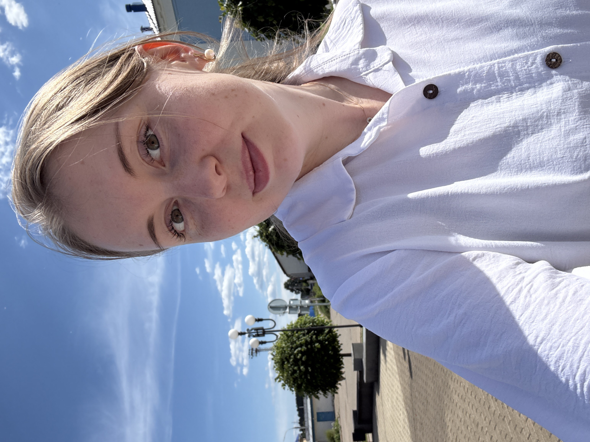 A fair-skinned young adult female is captured in a close-up, chest-up outdoor selfie. She has light brown hair, visible around her face with some strands swept back, and brown eyes. Freckles are present across her nose and cheeks. Her expression is a subtle, closed-mouth smile.
She is wearing a white, collared, long-sleeved button-up shirt crafted from a textured fabric, with two dark brown, circular, patterned buttons visible on the front placket. A slender, light-colored chain necklace is visible around her neck. She accessorizes with light-colored, spherical pearl-like earrings set in gold-toned hooks.
The scene is illuminated by bright natural sunlight originating from the upper right, creating distinct shadows on the left side of her body and shirt.
The background depicts an urban or semi-urban outdoor environment under a clear blue sky with scattered white clouds. To the left and slightly off-center, a black street lamp featuring three spherical white luminaires stands adjacent to a small, neatly trimmed spherical dark green tree or shrub. The ground beneath is paved with light brown or beige interlocking pavers. A low, grey concrete structure, possibly a bench or planter, is situated on the paved area to the left. In the mid-ground, a single-story white building with a blue door is partially visible to the far left. To the far right, a white building with a dark-colored roof and a black chimney is partially visible. Additional indistinct structures and foliage are discernible in the distant background. The combination of street furniture, paved areas, and architectural elements suggests a public square, plaza, or a wide thoroughfare within an unidentified town or city.